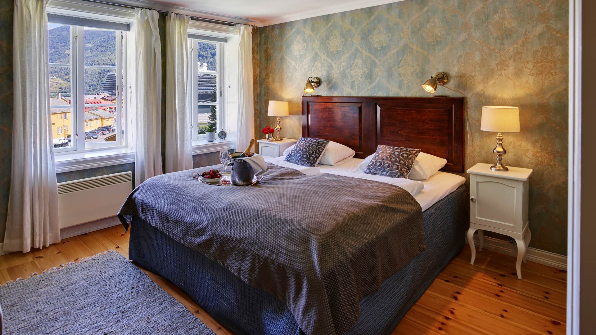 Historisk hotellrom med antikkinspirert grønn tapet, vindu med utsikt mot Flåm og dobbeltseng med et brett med champagne og jordbær på. 