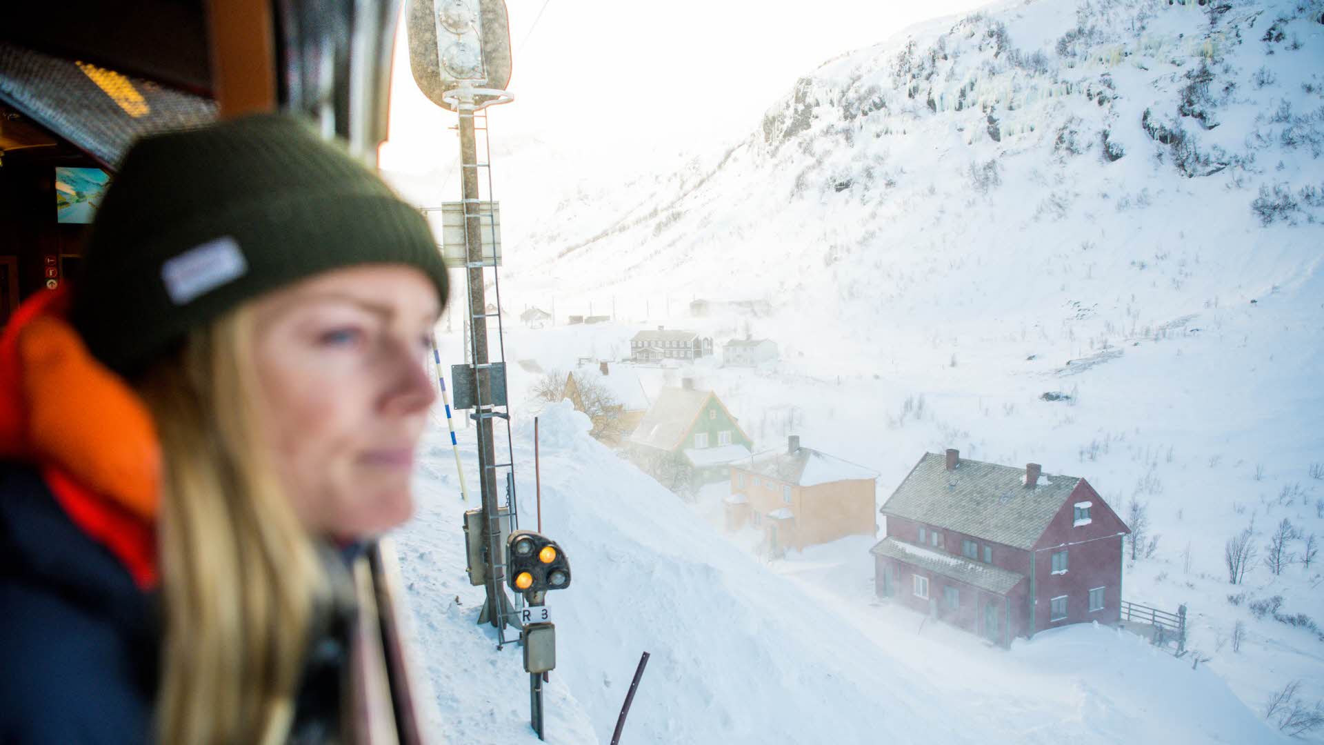 Una mujer con un sombrero mirando por la ventana del tren de Flåm, cerca de unas casas cubiertas de nieve.