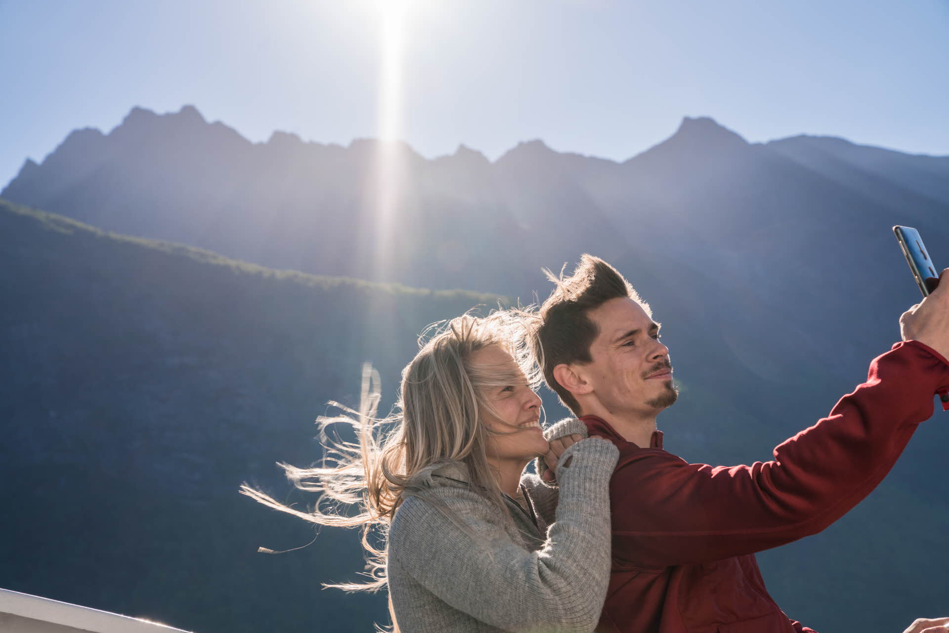 Young happy couple taking selfie onboard fjord cruise Hjorundfjord. Tall spiky wild mountains in background sunny day