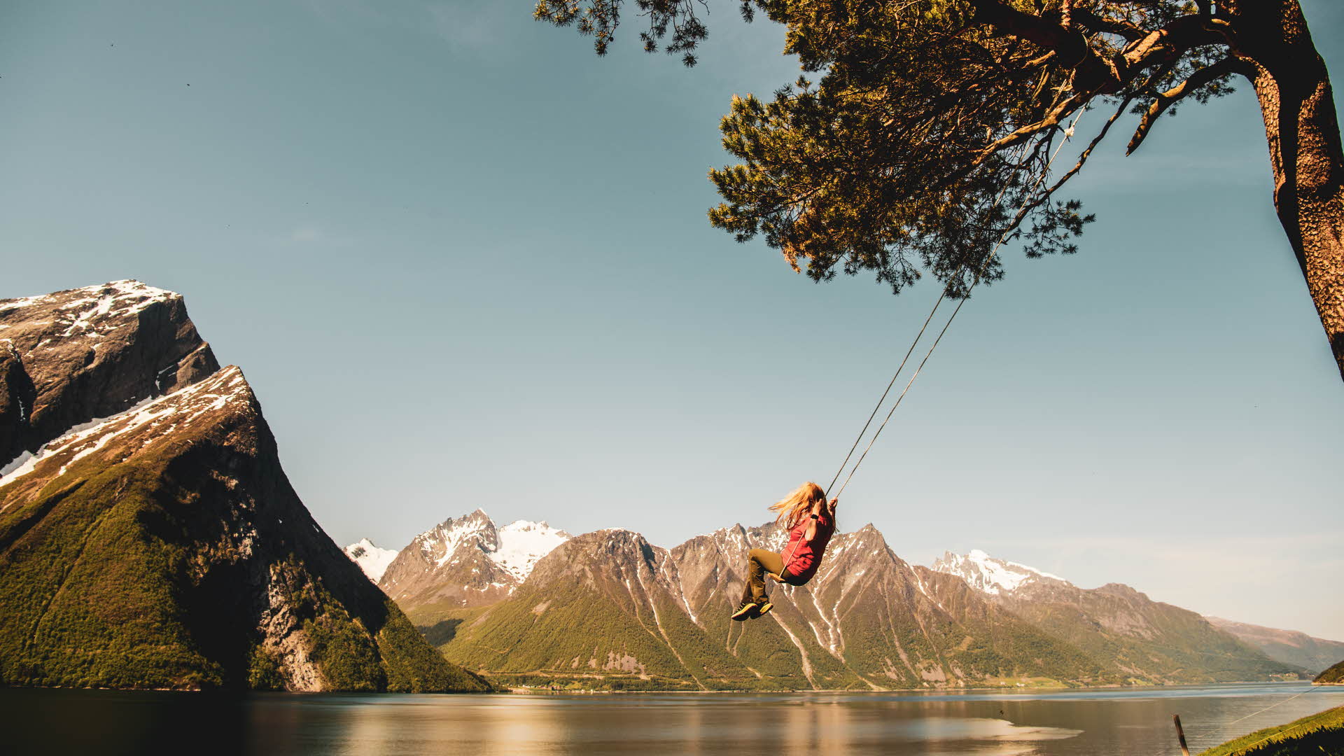 Woman enjoying tree-swing at Trandal farm by Hjorundfjord. Dramatic alpine mountains with snow-capped mountains