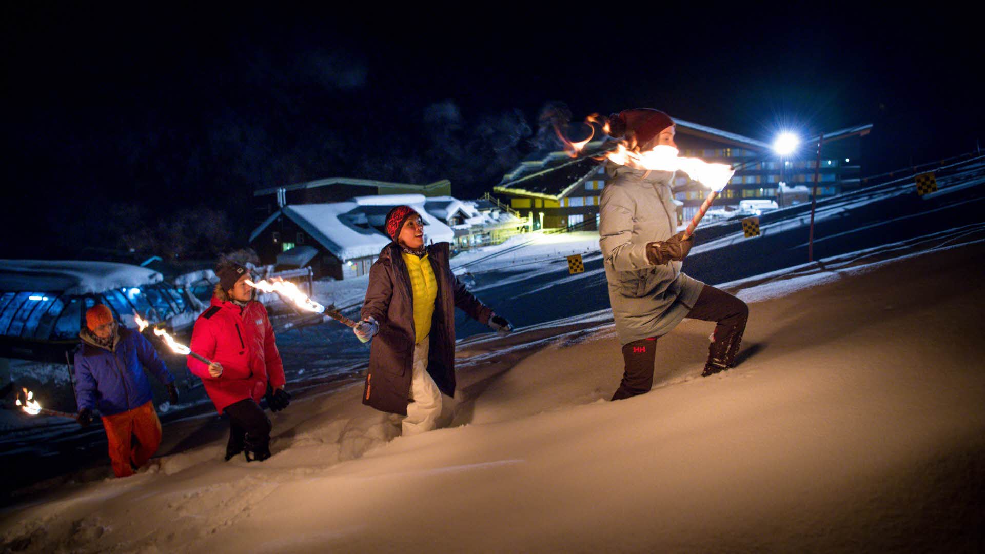 Devant la station de ski de Myrkdalen, 4 personnes marchent sur une colline escarpée la nuit en tenant des torches allumées dans leurs mains. La neige est profonde.