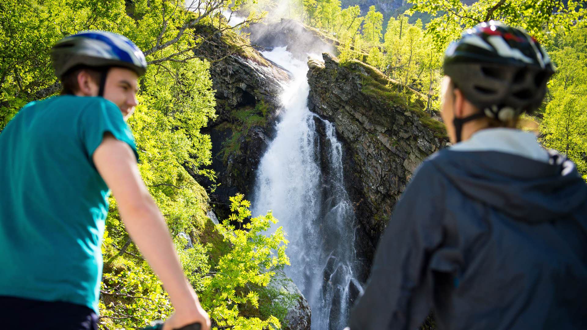 Two people wearing bike helmets and looking towards a waterfall in Flåmsdalen