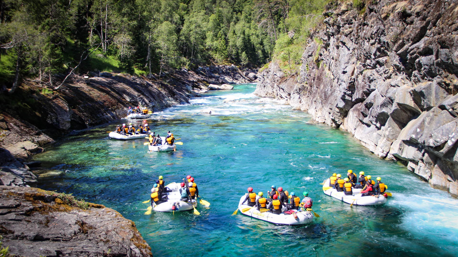 Six rafts full of people in life jackets on a clear river in a canyon.