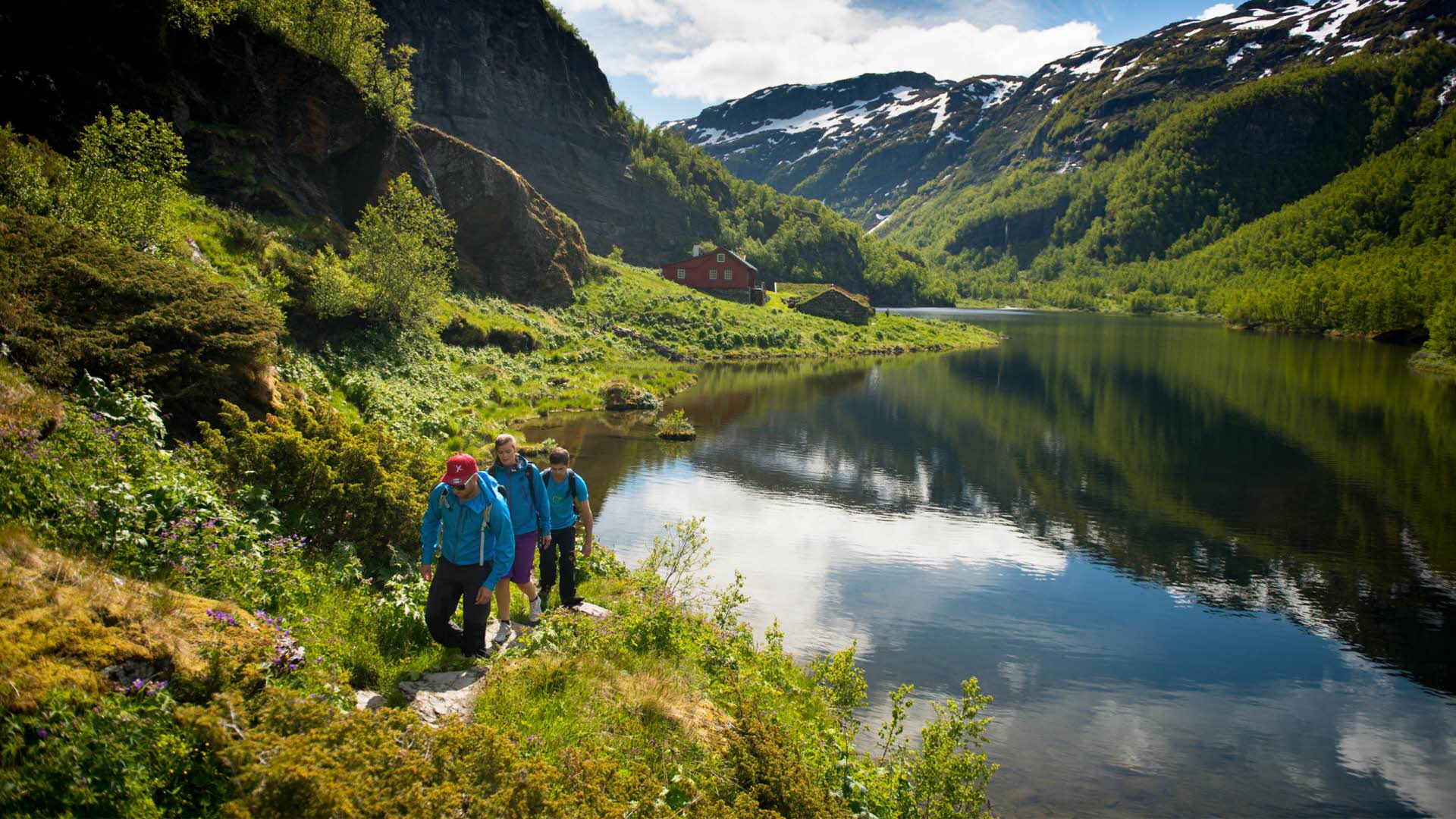 Groupe de trois personnes marchant sur un chemin longeant un lac entouré de montagnes verdoyantes parsemées de neige. 