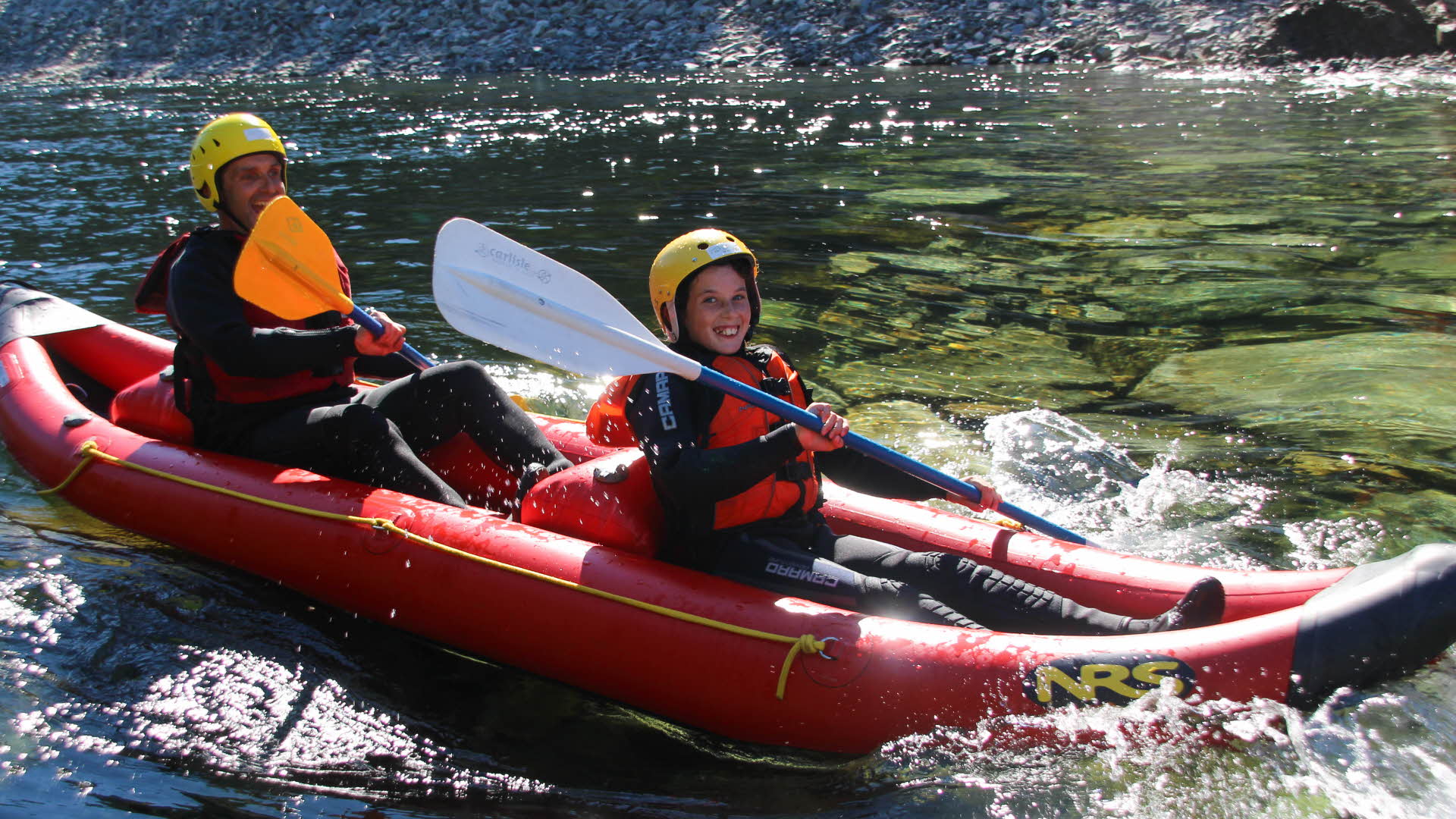 两个男孩正在一条清澈的河流上的红色橡皮船中微笑。手里拿着桨，身上穿着救生衣和防寒泳衣，头上戴着头盔。