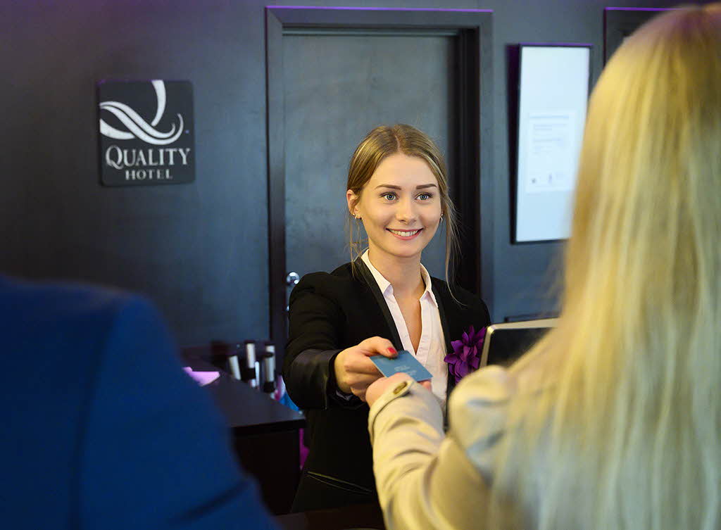 A receptionist handing a key to a blond woman seen from behind. 