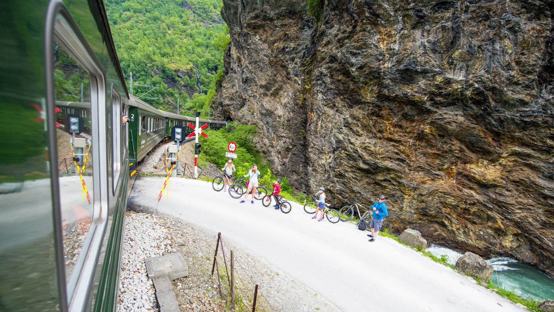 Family of 4 on bicycles waiting when Flam Railway is passing the Navvy road in Flåm valley with green river flowing behind