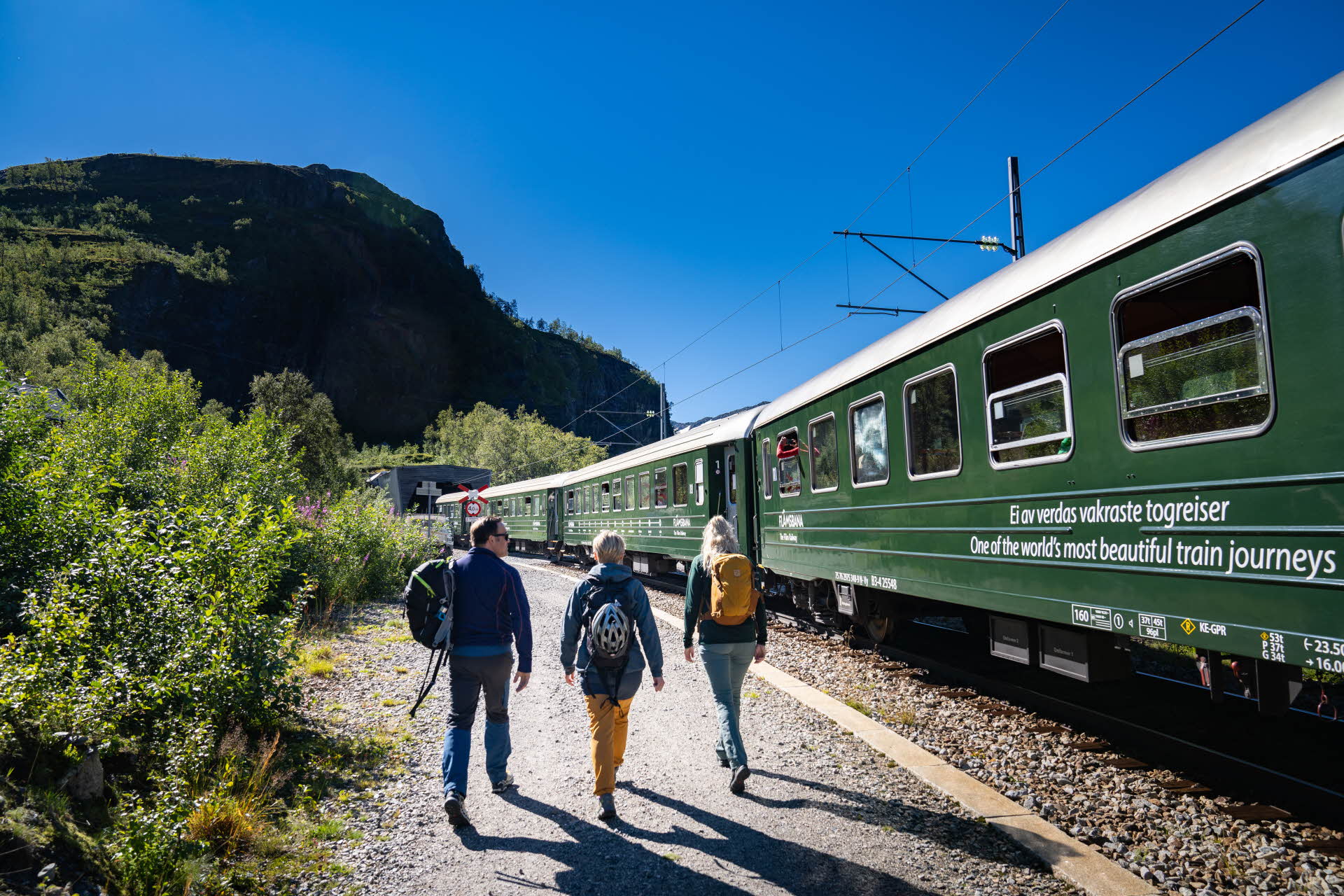 Tres personas caminan a lo largo del ferrocarril de Flåm, un glorioso día de verano. 