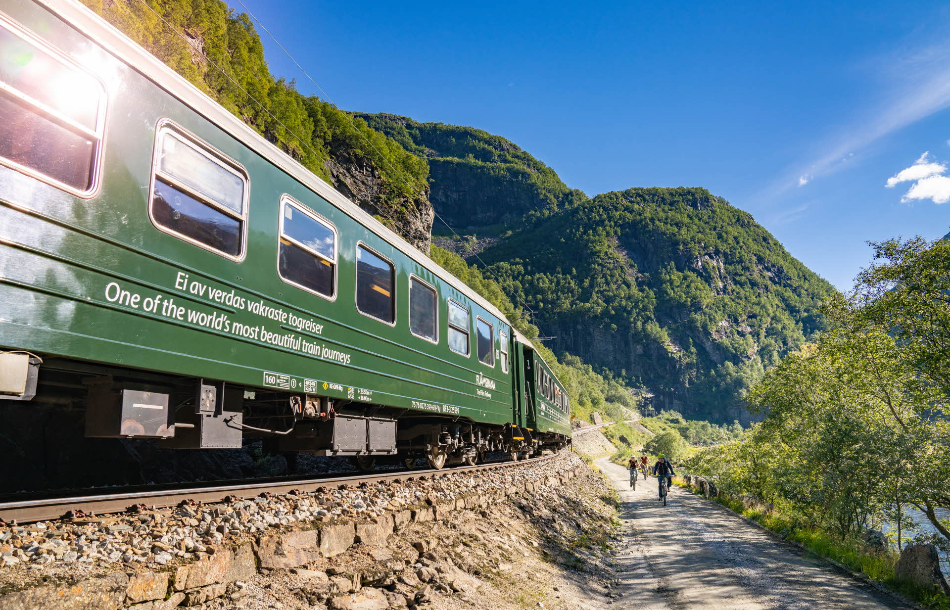 Cyclists on a gravel road next to the green Flåm Railway train. Mountains covered in forest.