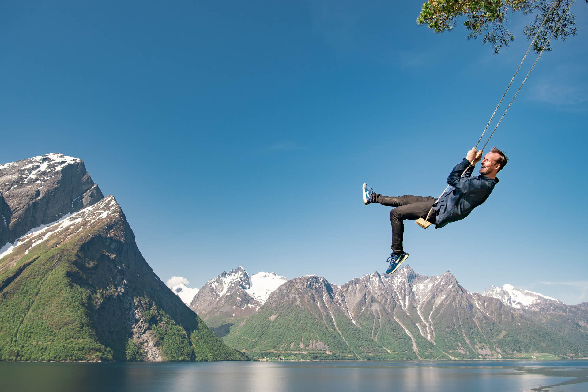 A man on a swing above the Hjørundfjord. The Sunnmøre Alps with patches of snow in the foreground.