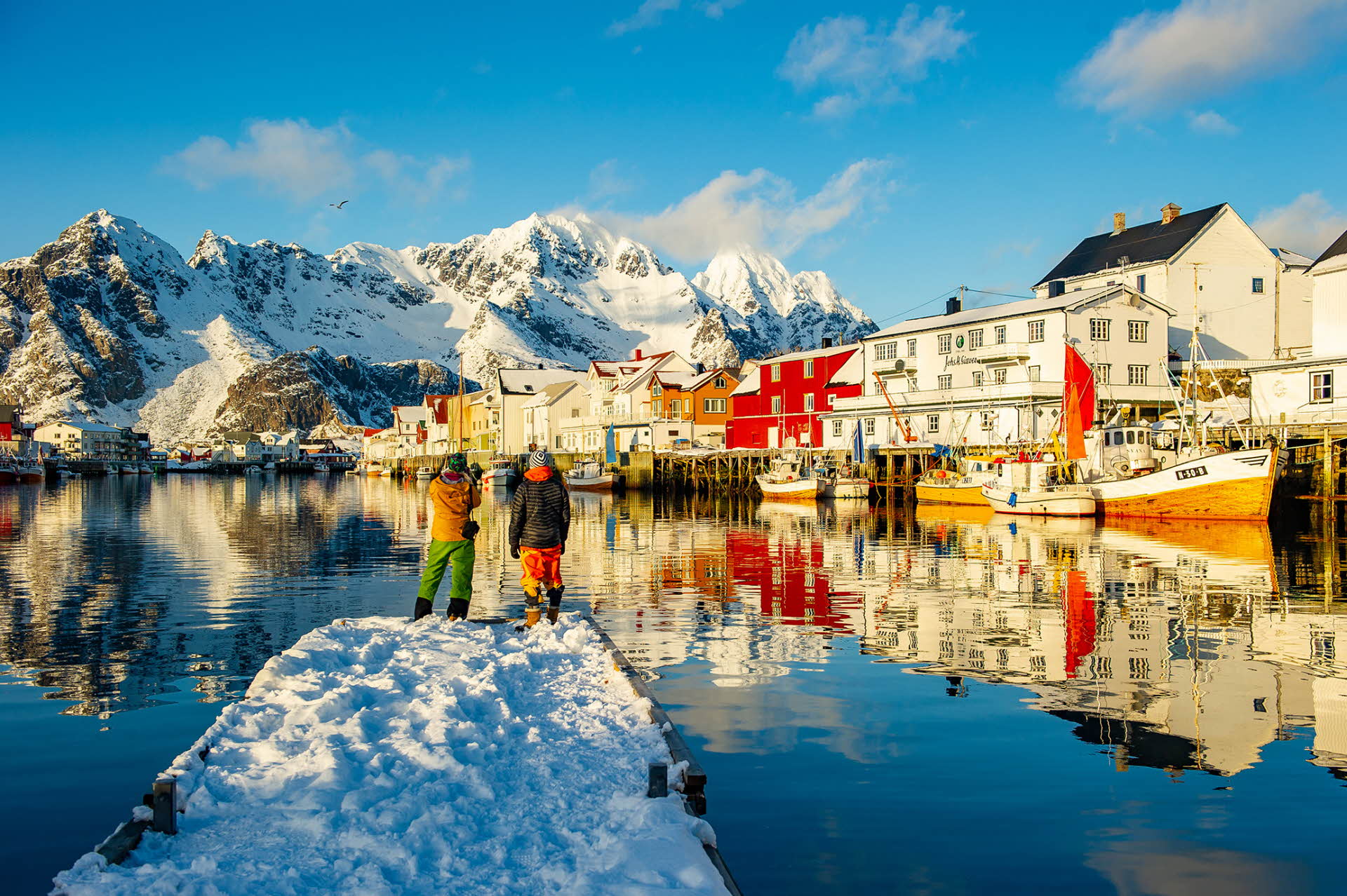 Two men standing on a quay in winter dress looking at a Lofoten Island village. Snowy mountains in the foreground.