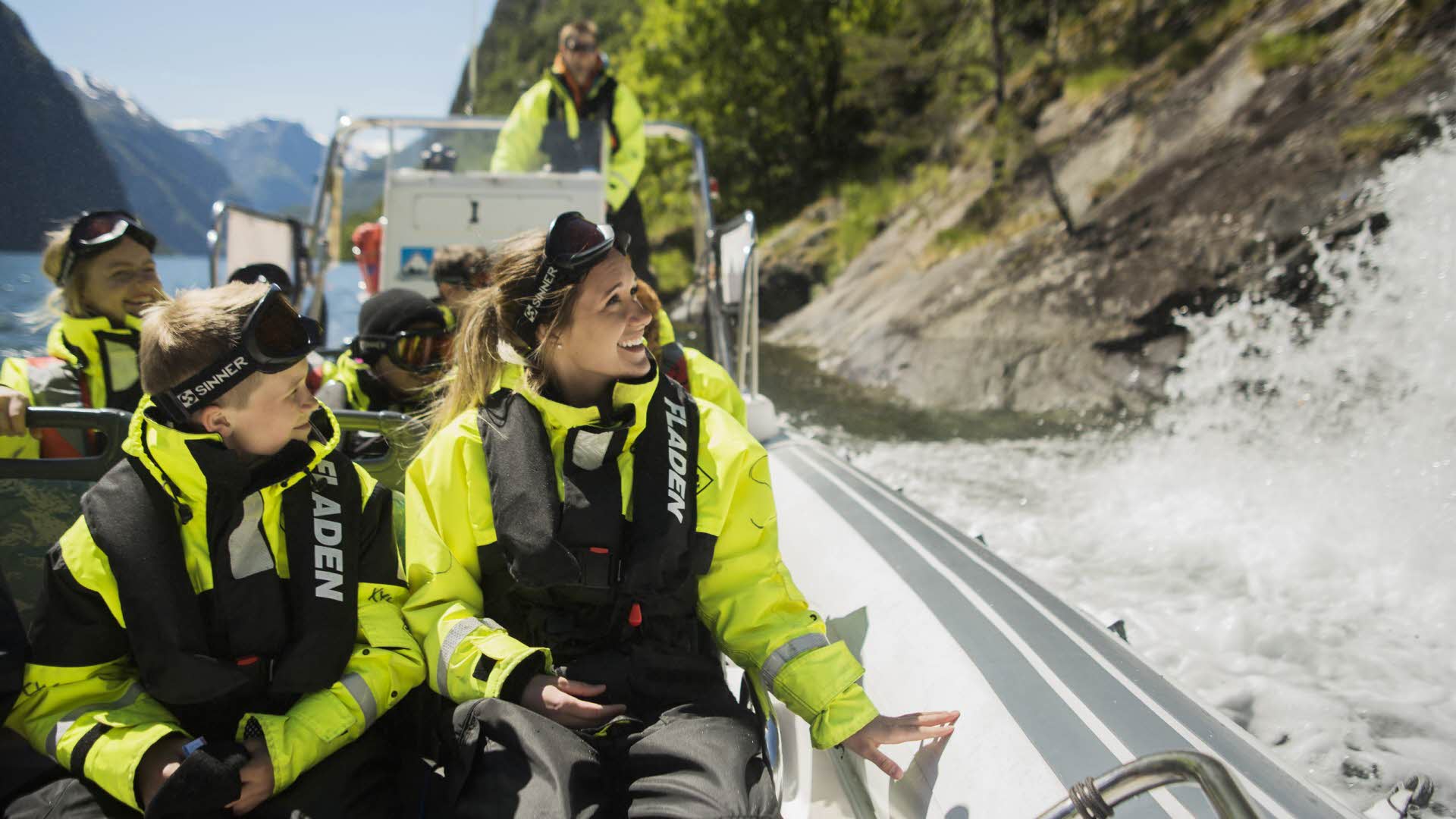 Una mujer y unos niños sonriendo con trajes secos en un bote junto a una cascada del Nærøyfjord