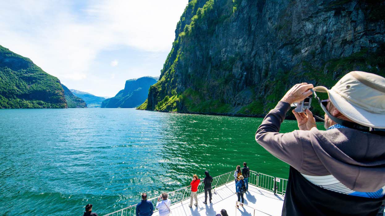 En mann med hatt tar bilde fra øvre dekk på båten på Nærøyfjorden, mens andre nyter utsikten nedenfor.