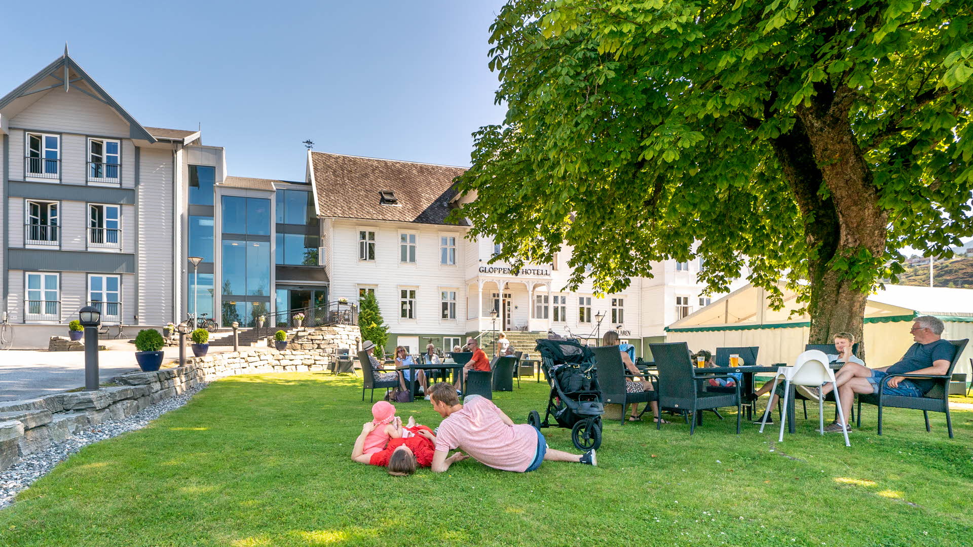 Blick auf das Gloppen Hotell vom Garten aus. Eine Familie liegt im Gras, andere Gäste sitzen an Gartentischen. 