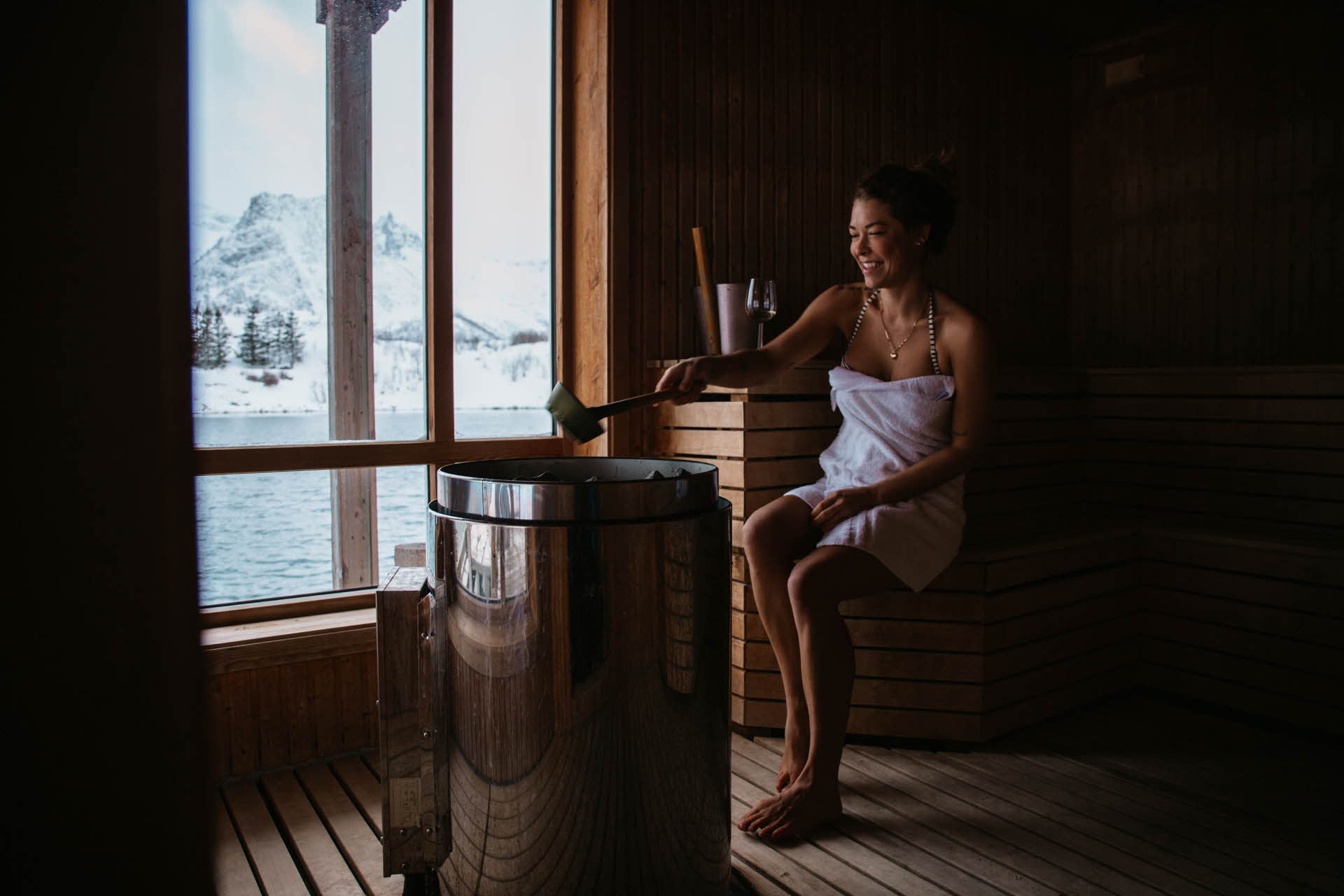 A woman wearing a towel in a sauna by large windows. Snowy mountains outside 