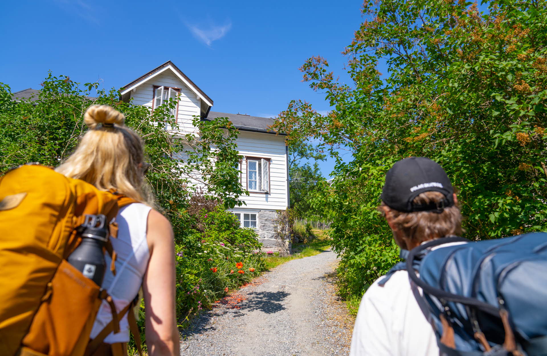 A man and a woman wearing backpacks looking at an old house in Geiranger