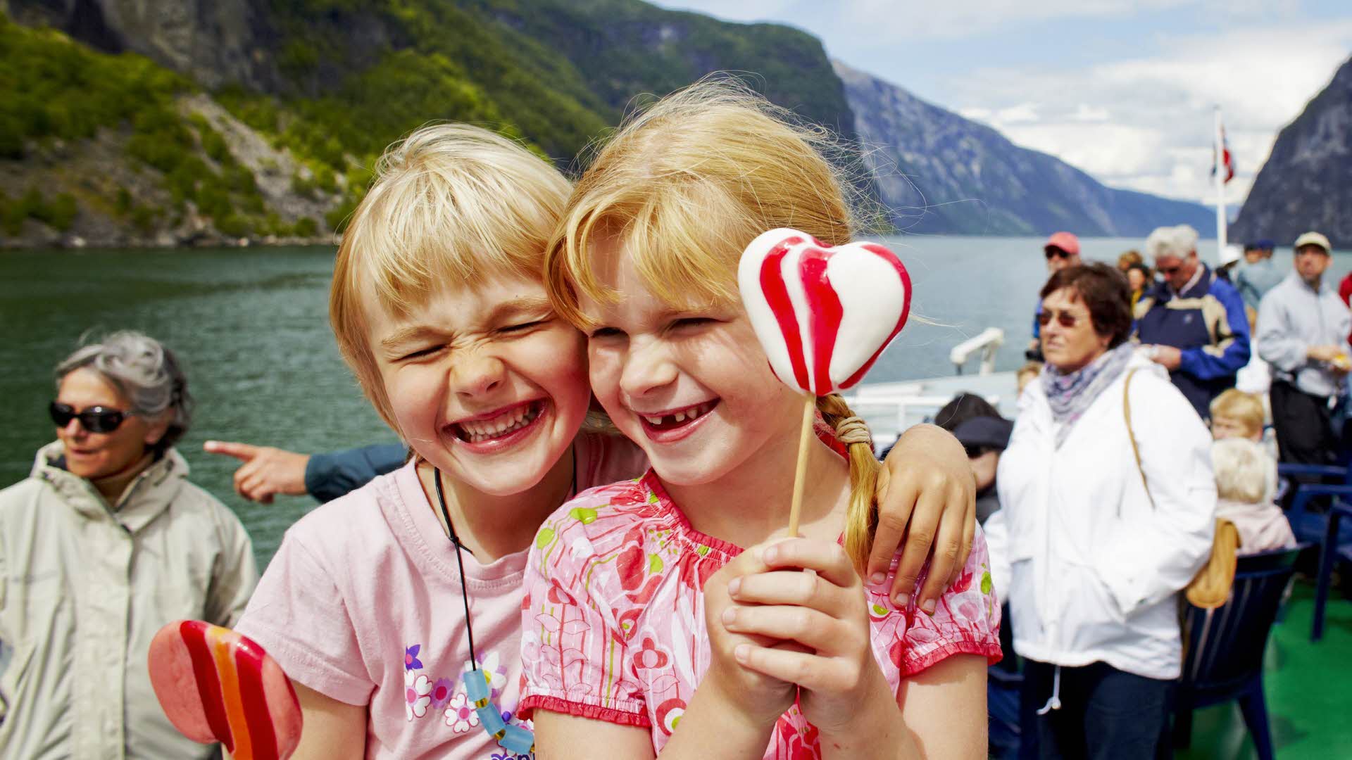 两个兴高采烈的女孩正在经典的游船上品味棒棒糖；游船正航行在纳柔依峡湾 