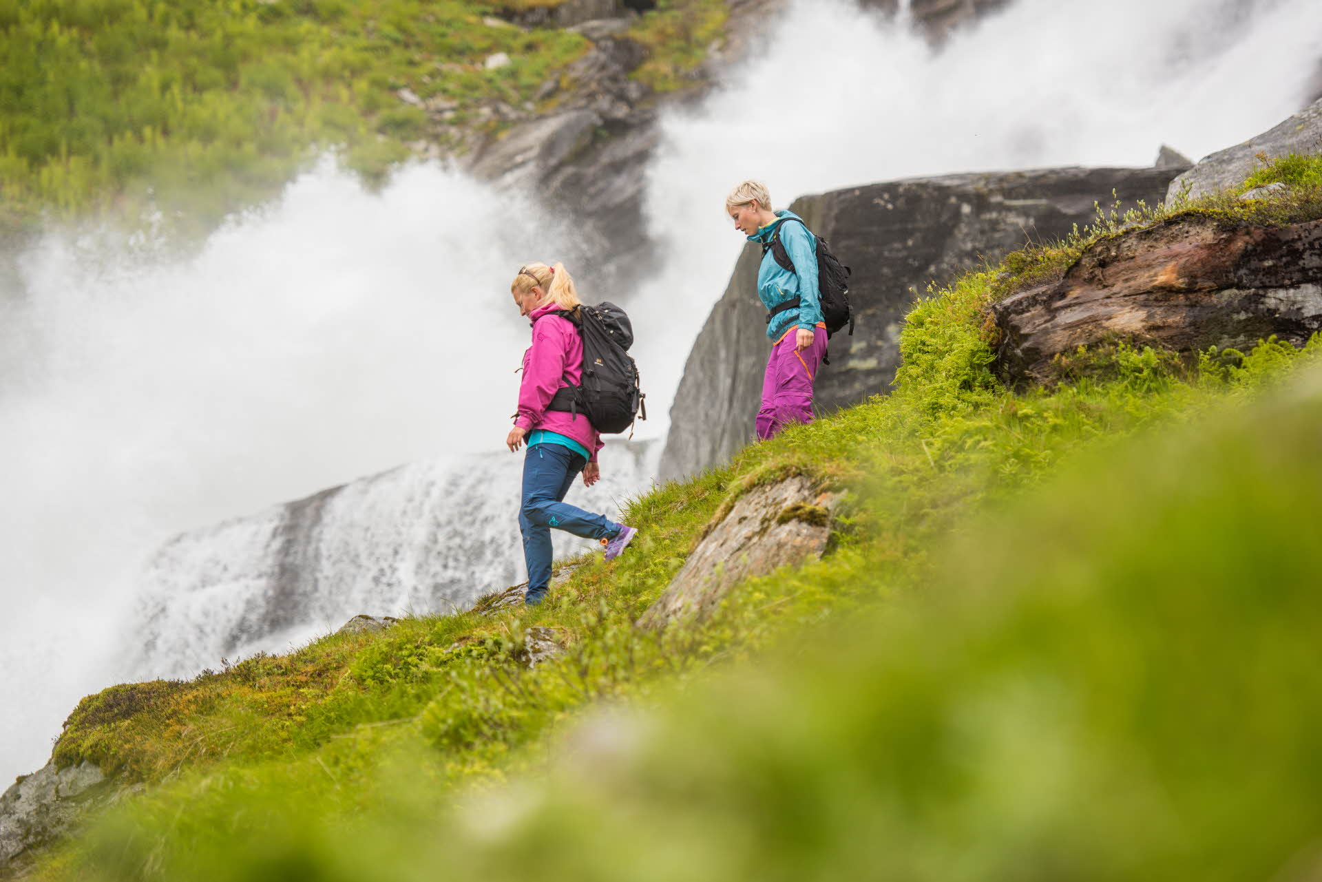 Two women wearing bright coloured hiking clothes walking near a large waterfall