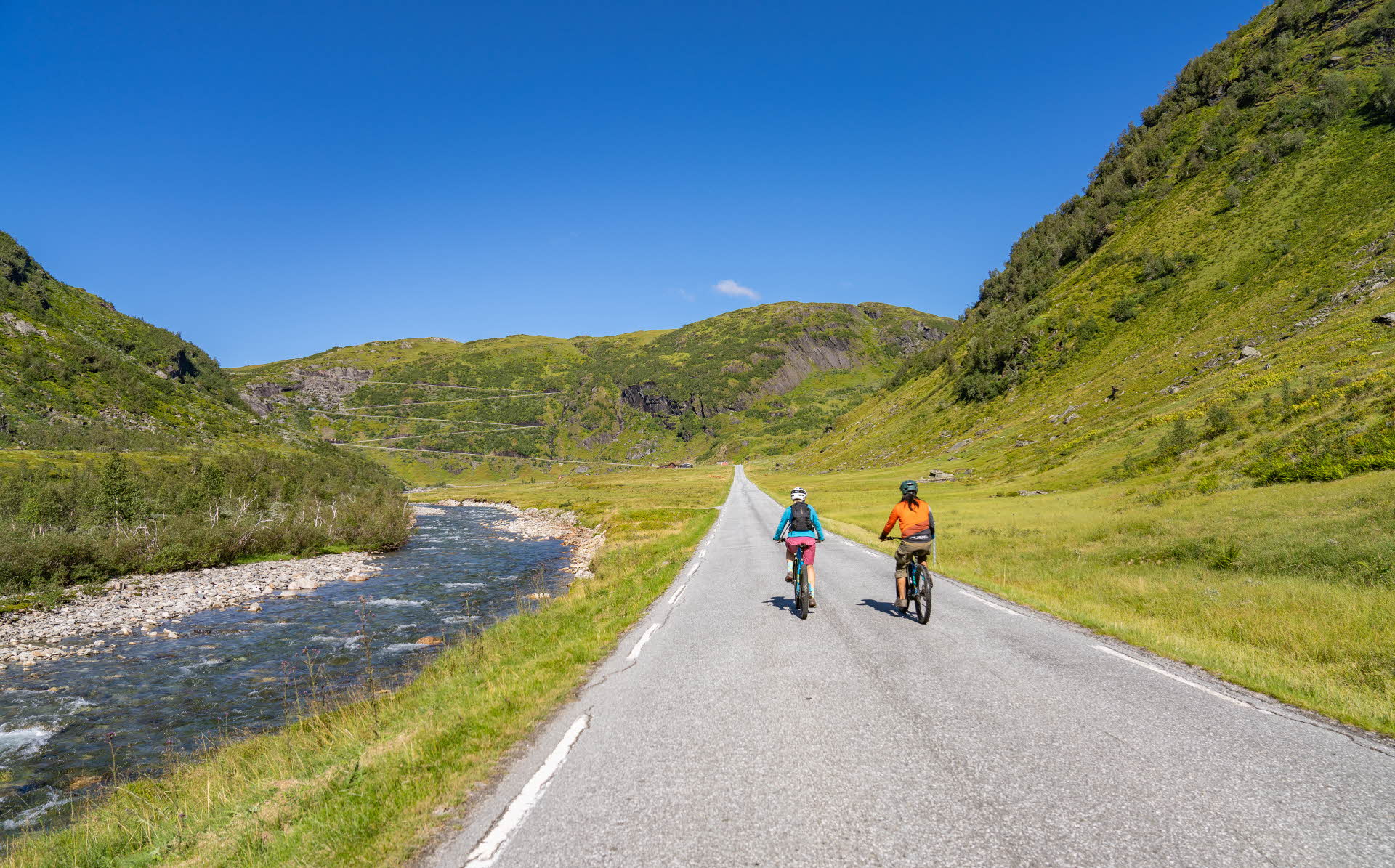 Two cyclists on a road by a river and steep, green mountains on every side.