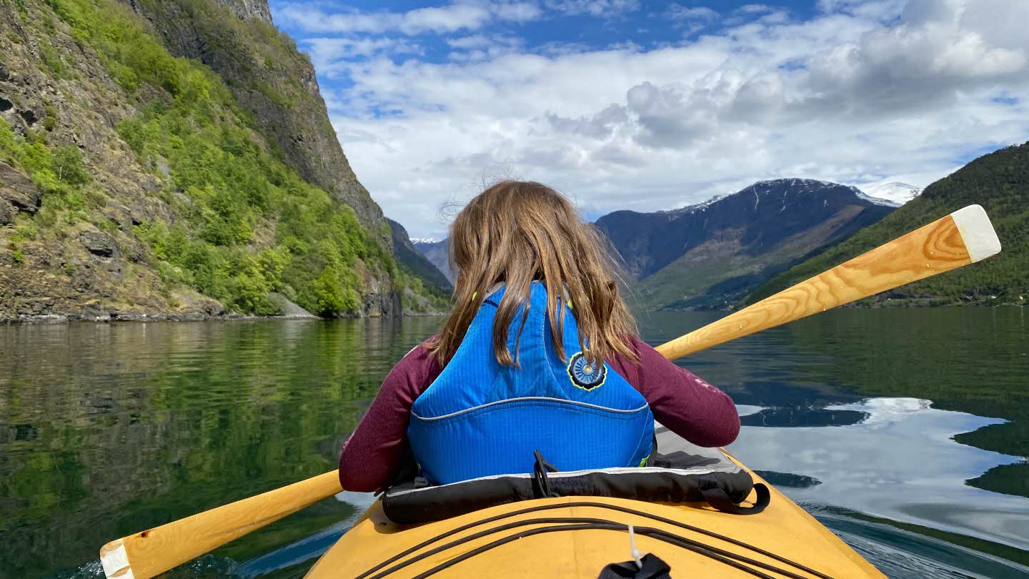 A child in a blue life vest sitting in the front of a yellow double kayak on the Aurlandsfjord. 