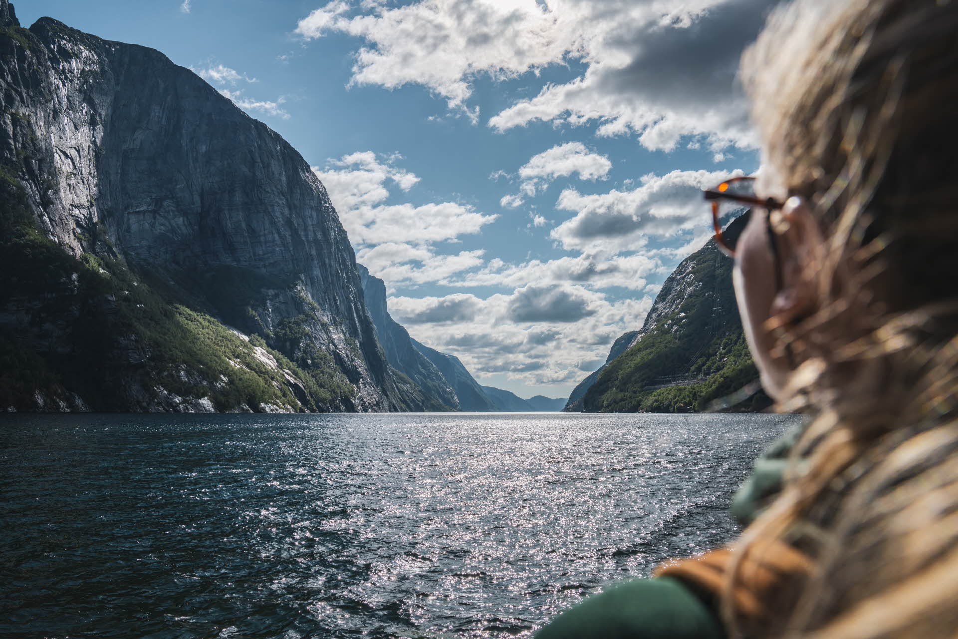 前景中一个戴着太阳镜的金发女人看向吕瑟峡湾 (Lysefjord) 和绵延的山脉