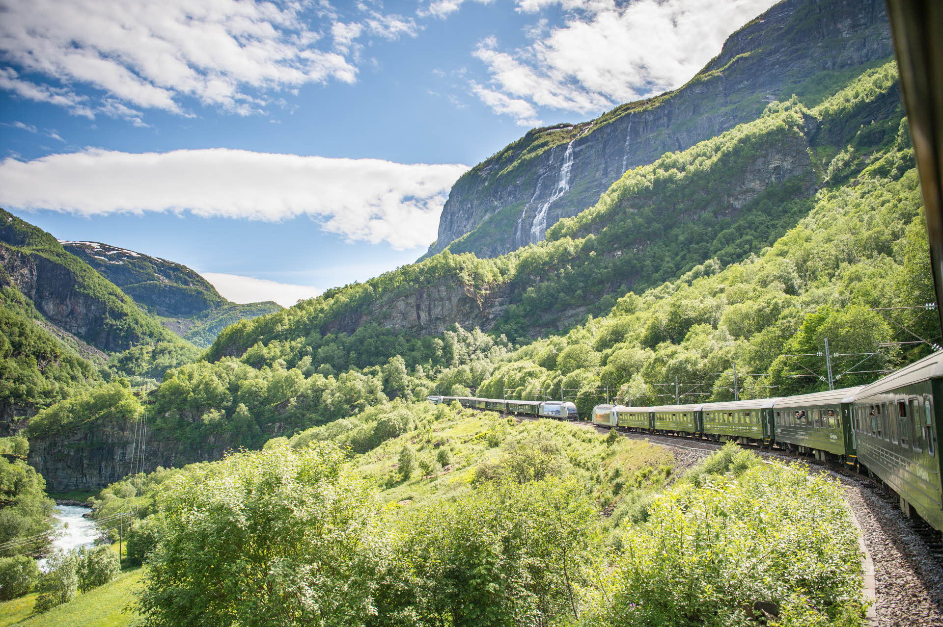 El valle de Flåm y dos trenes encontrándose, vistos desde la ventana de un tren. Unas laderas verdes, el cielo azul y una cascada. 