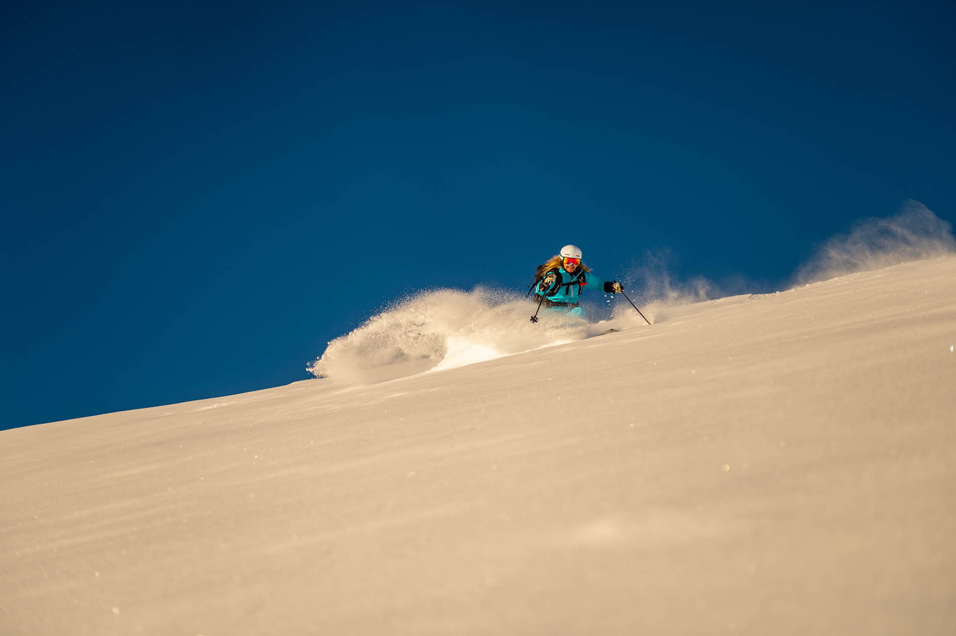 A woman in a blue jacket and white helmet skiing in fresh powder snow. Blue sky in the background. 