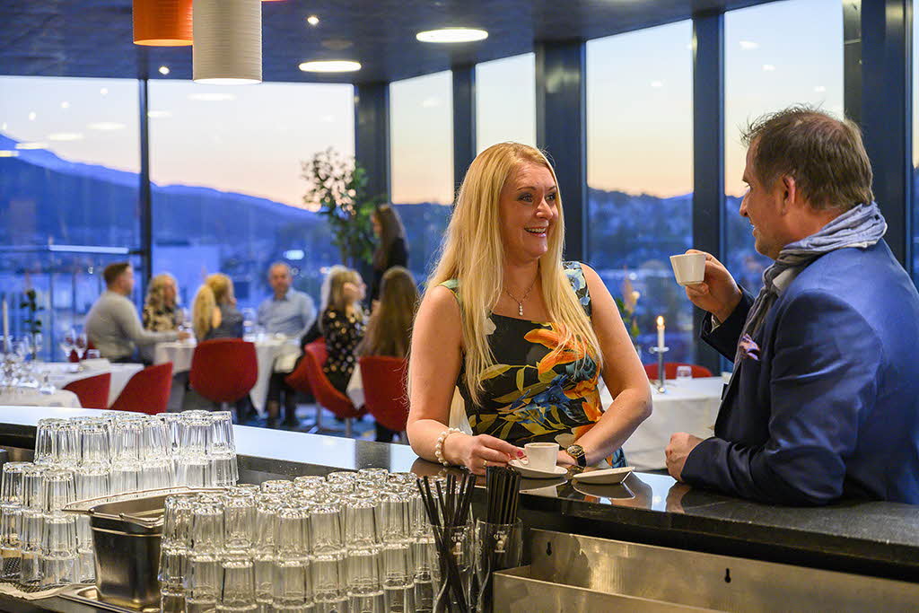 En mann og en kvinne drikker kaffe og snakker ved baren på Linken. Folk rundt bord og utsikt over Narvik bak.