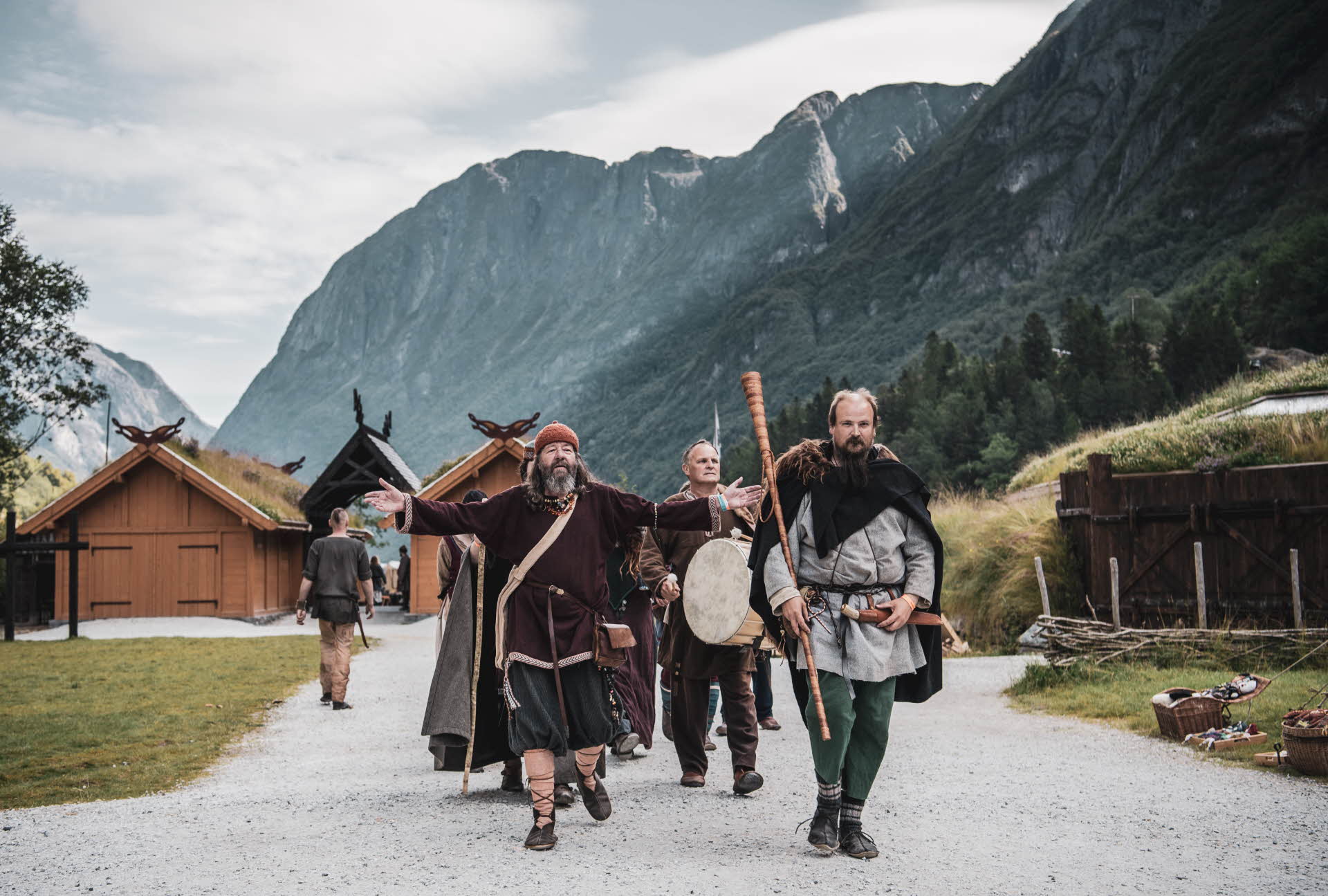 Eine Gruppe männlicher Wikinger läuft mit Instrumenten und anderen Accessoires ins Wikingerdorf hinein. Im Hintergrund sind hohe Berge zu sehen.