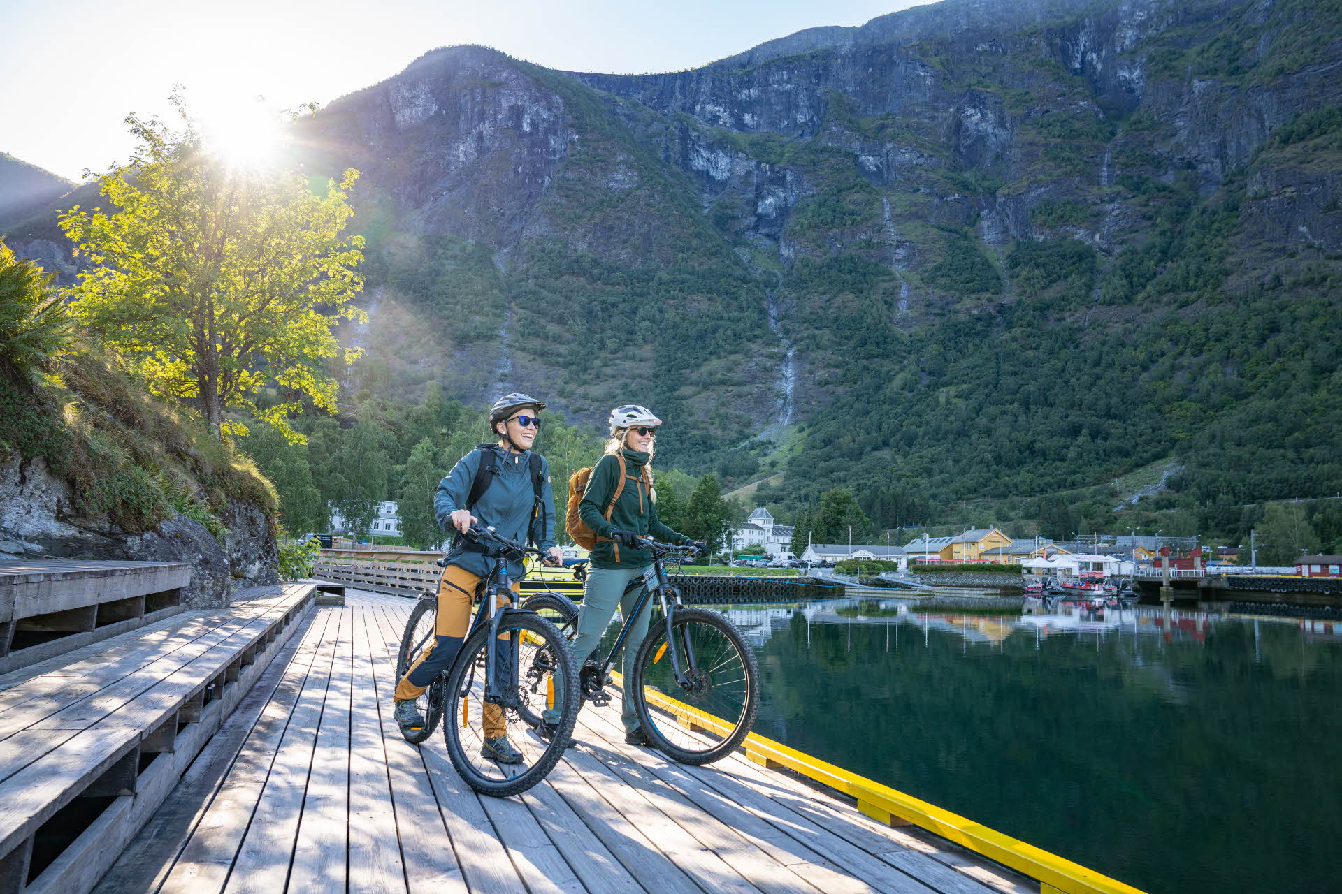 To kvinner står med sykler på en brygge og ser ut mot fjorden. Flåm og fjell bak dem.
