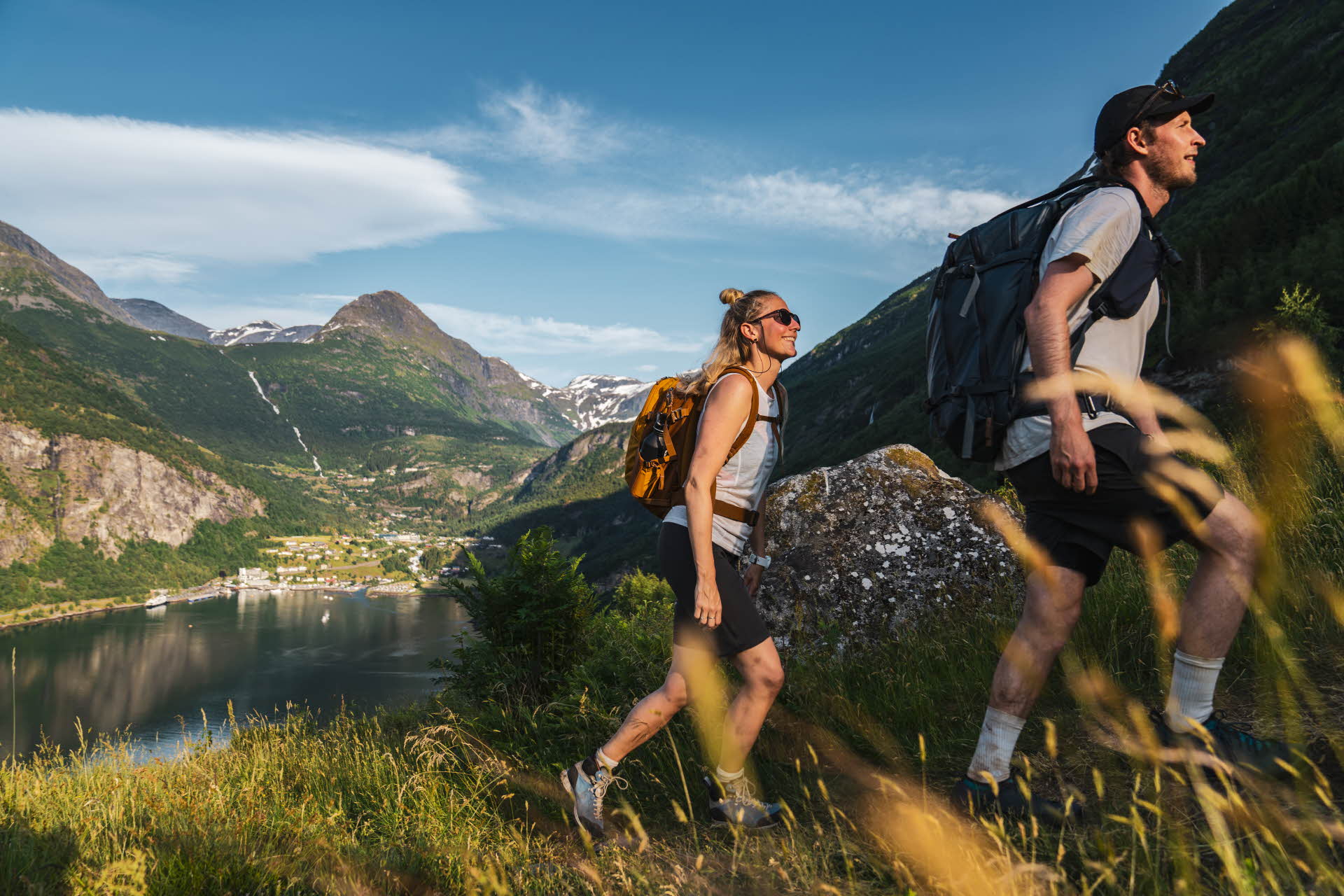 Un homme et une femme gravissant une colline, avec Geiranger et le Geirangerfjord à l’arrière-plan.