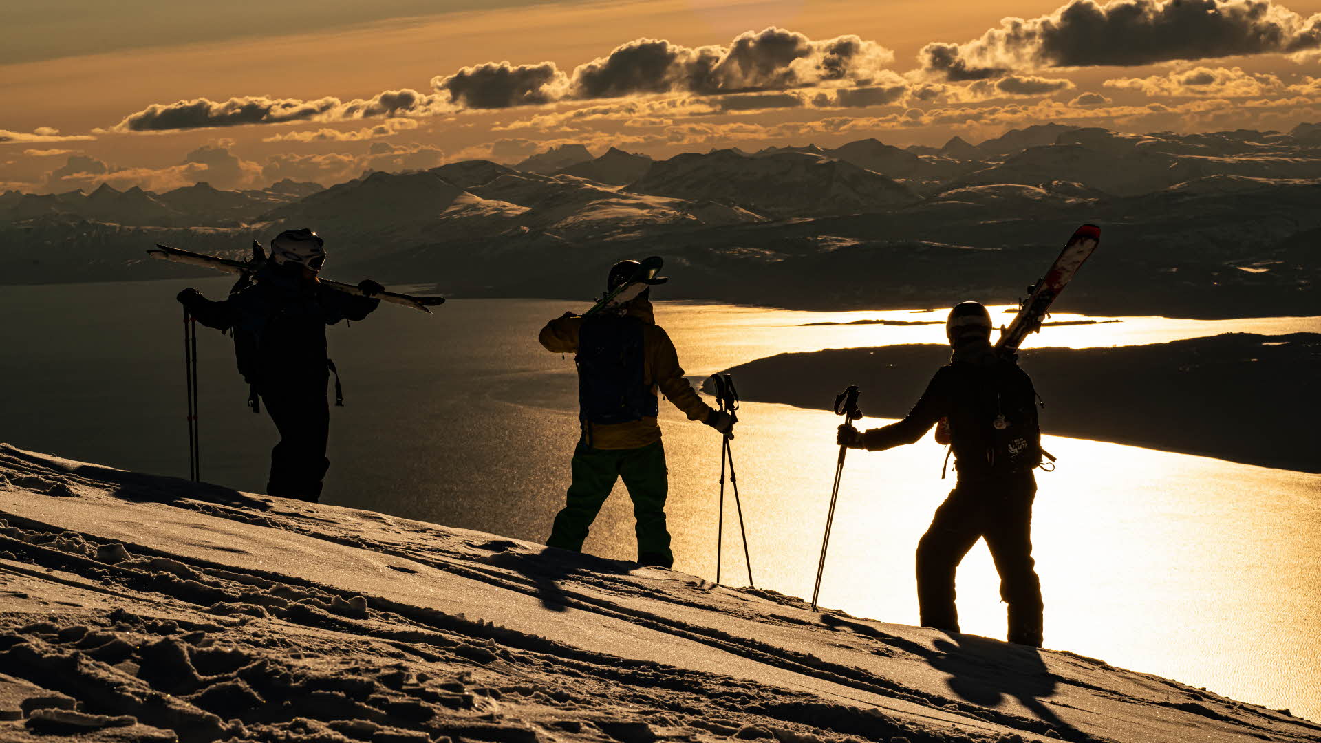 スキー板を肩にかつぎ、オーフォートフィヨルドを見つめる３人のスキーヤー。