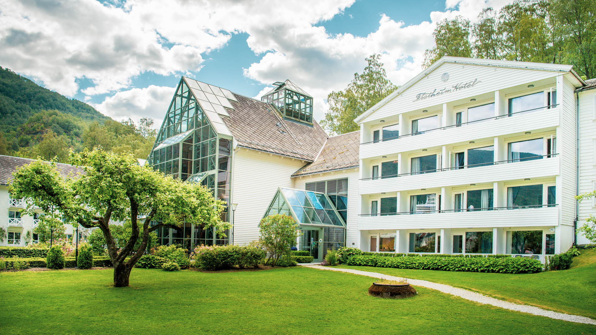 Außenansicht des Hotels Fretheim in Flåm mit grünen Bäumen, blühenden Büschen und gepflegtem Rasen