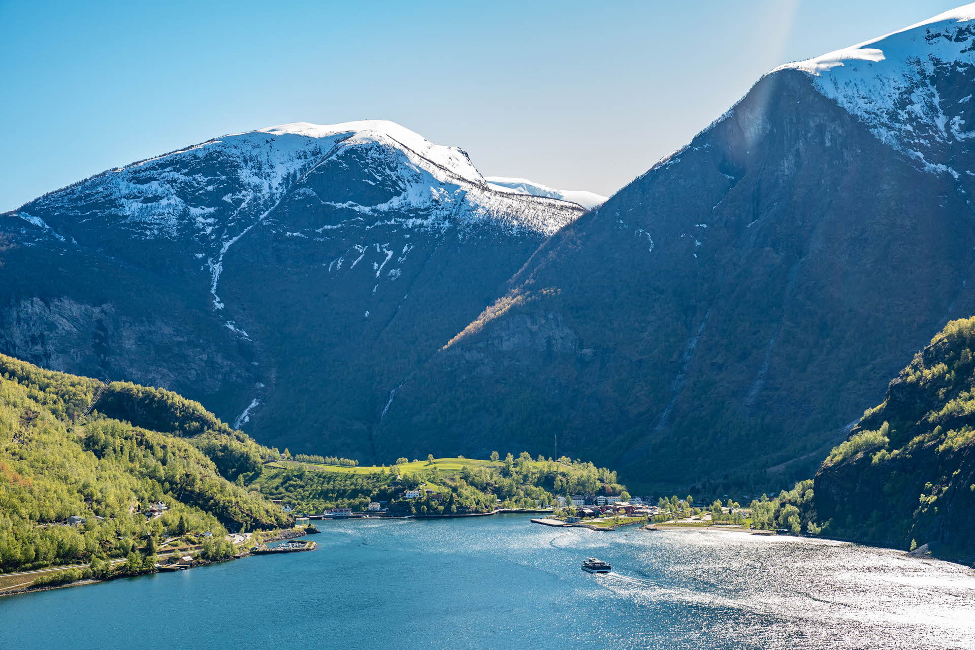 Flåm mit schneebedeckten Bergen im Hintergrund und einem Schiff, das auf dem Fjord unterwegs ist
