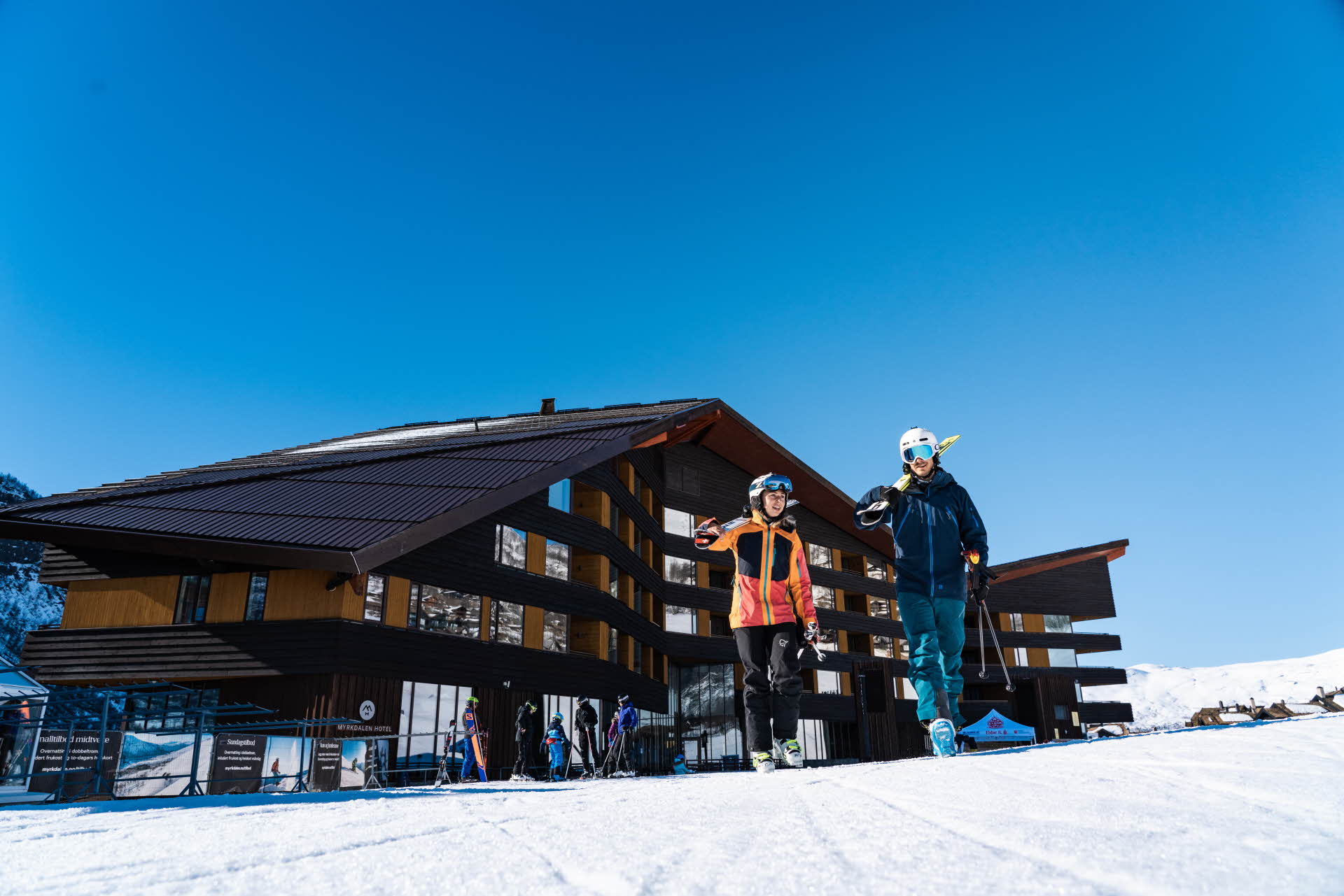 一男一女戴着滑雪头盔，拿着滑雪板，走在迈克达林酒店 (Myrkdalen Hotel) 旁边的雪地上