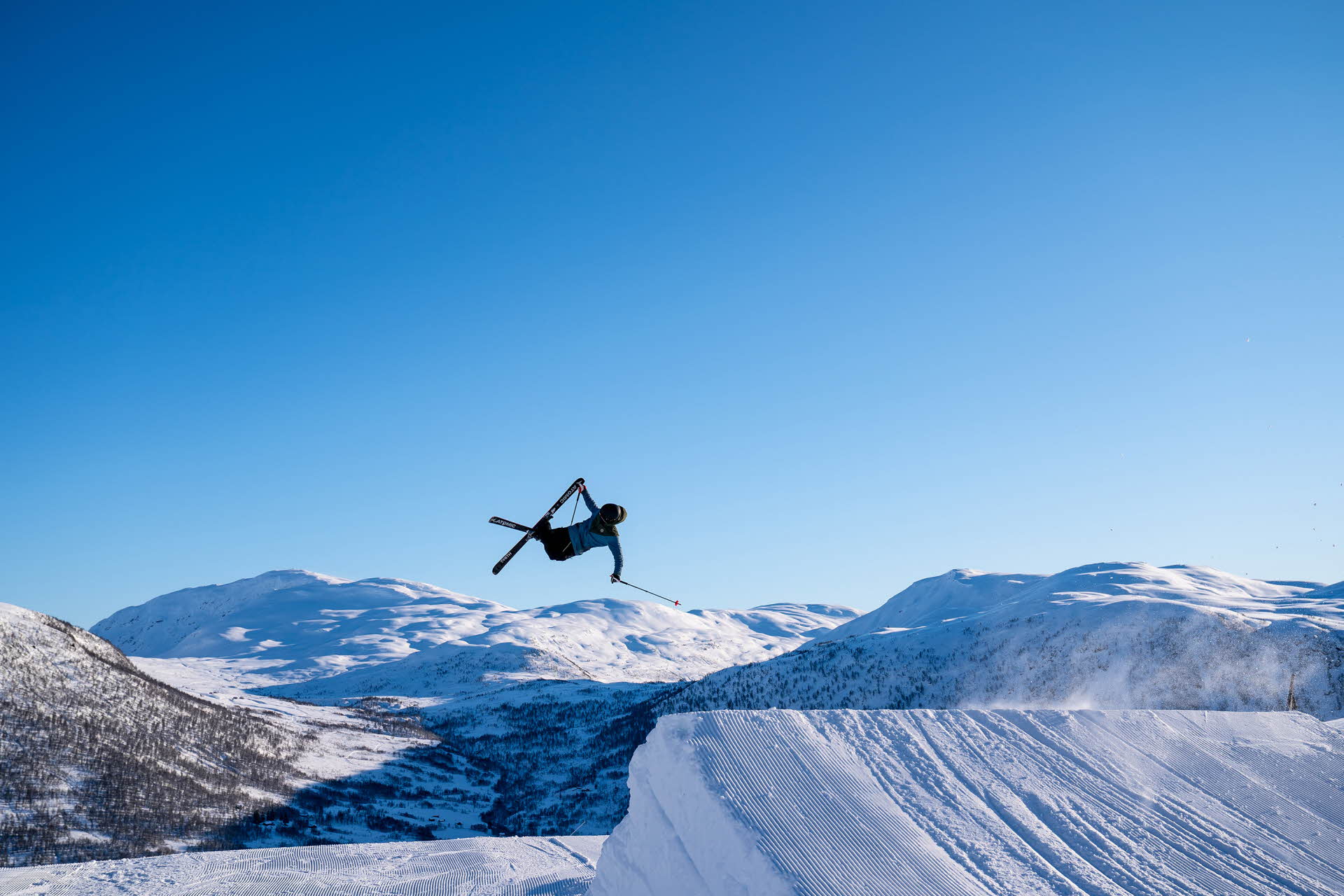Una persona saltando con los esquís en Myrkdalen Mountain Resorts. Bajo un cielo azul y rodeado por un paisaje de montaña. 