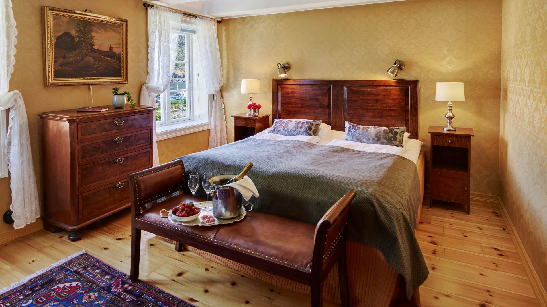 Historisk rom på Fretheim Hotel med dobbeltseng, blondegardiner og et brett med champagne og jordbær oppå en antikk benk i skinn