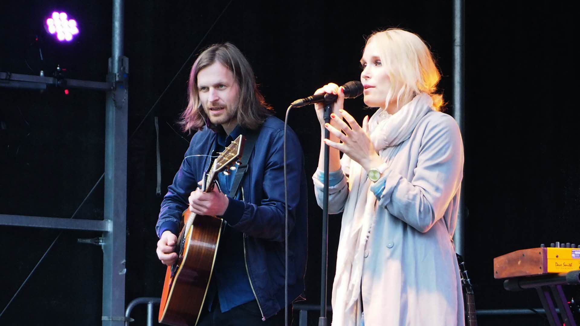 Une femme blonde chantant dans un micro et un homme aux cheveux longs jouant de la guitare sur scène.