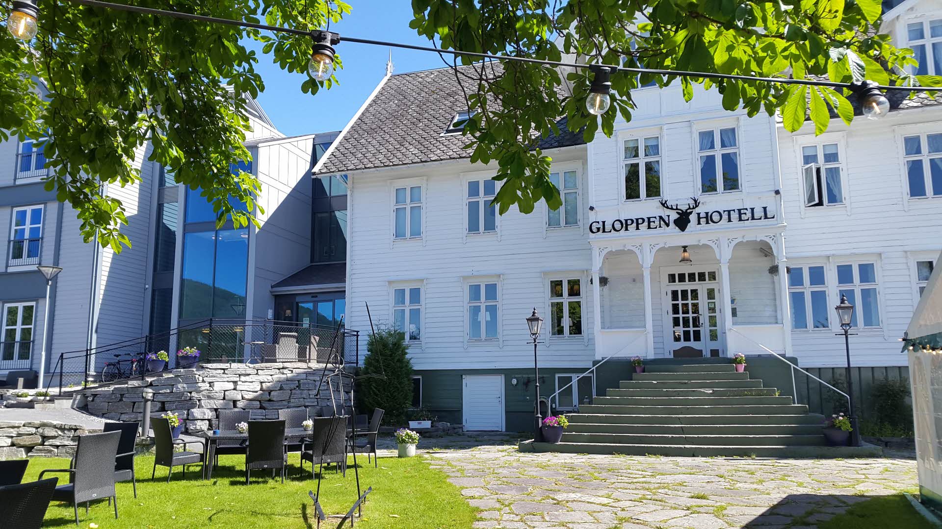 Foto exterior del Gloppen Hotell en un día de verano con sillas en el jardín y enmarcado en hojas verdes.