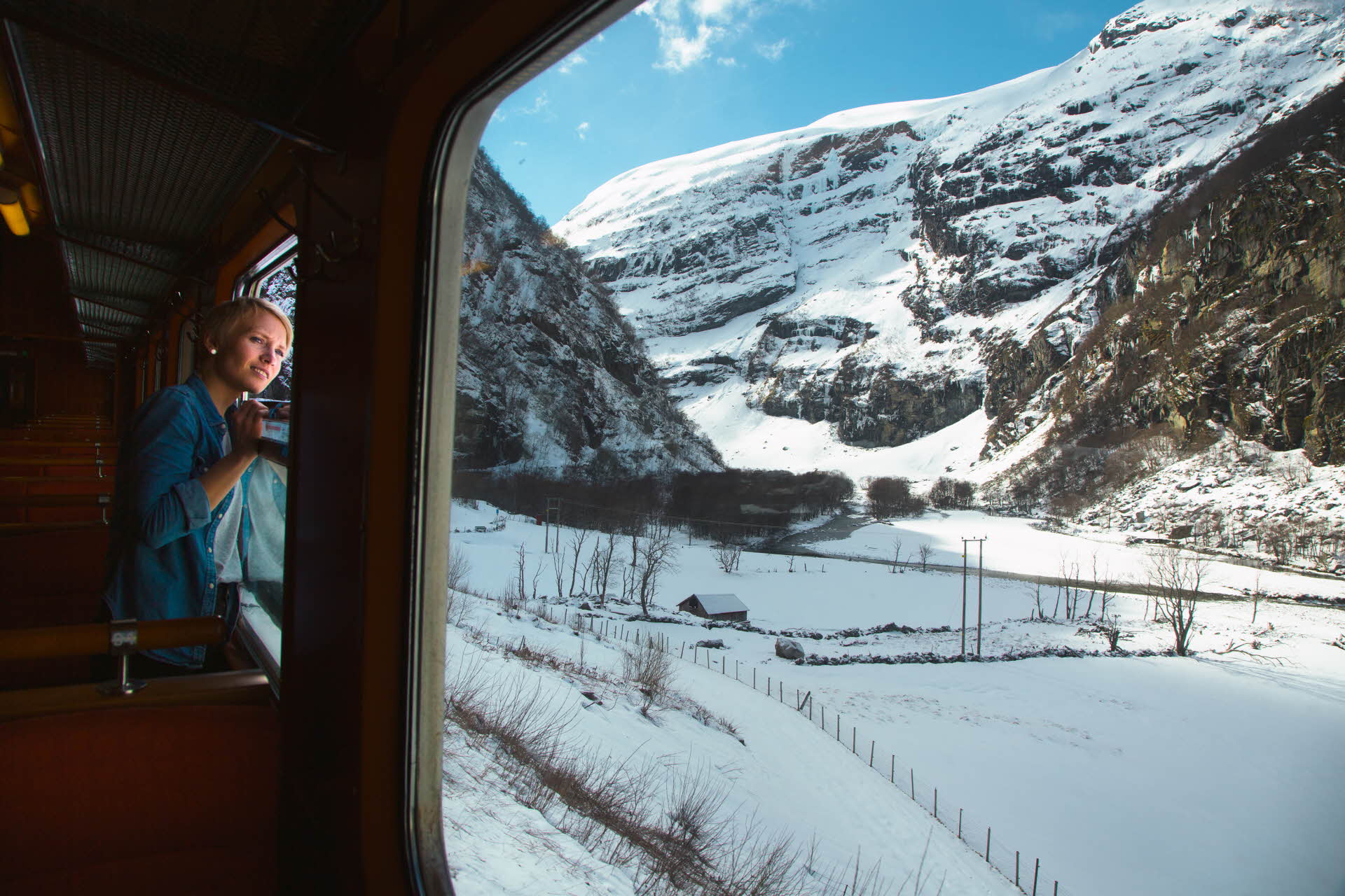 A woman looking out of a window from inside the Flåm Railway. Snowy and mountainous landscape outside. 