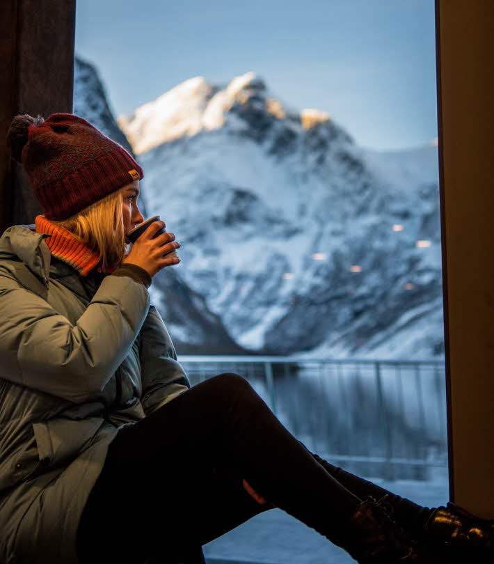 Woman in winter clothing with hot drink sitting in window post onboard Vision of The Fjords enjoying the UNESCO fjord landscape 