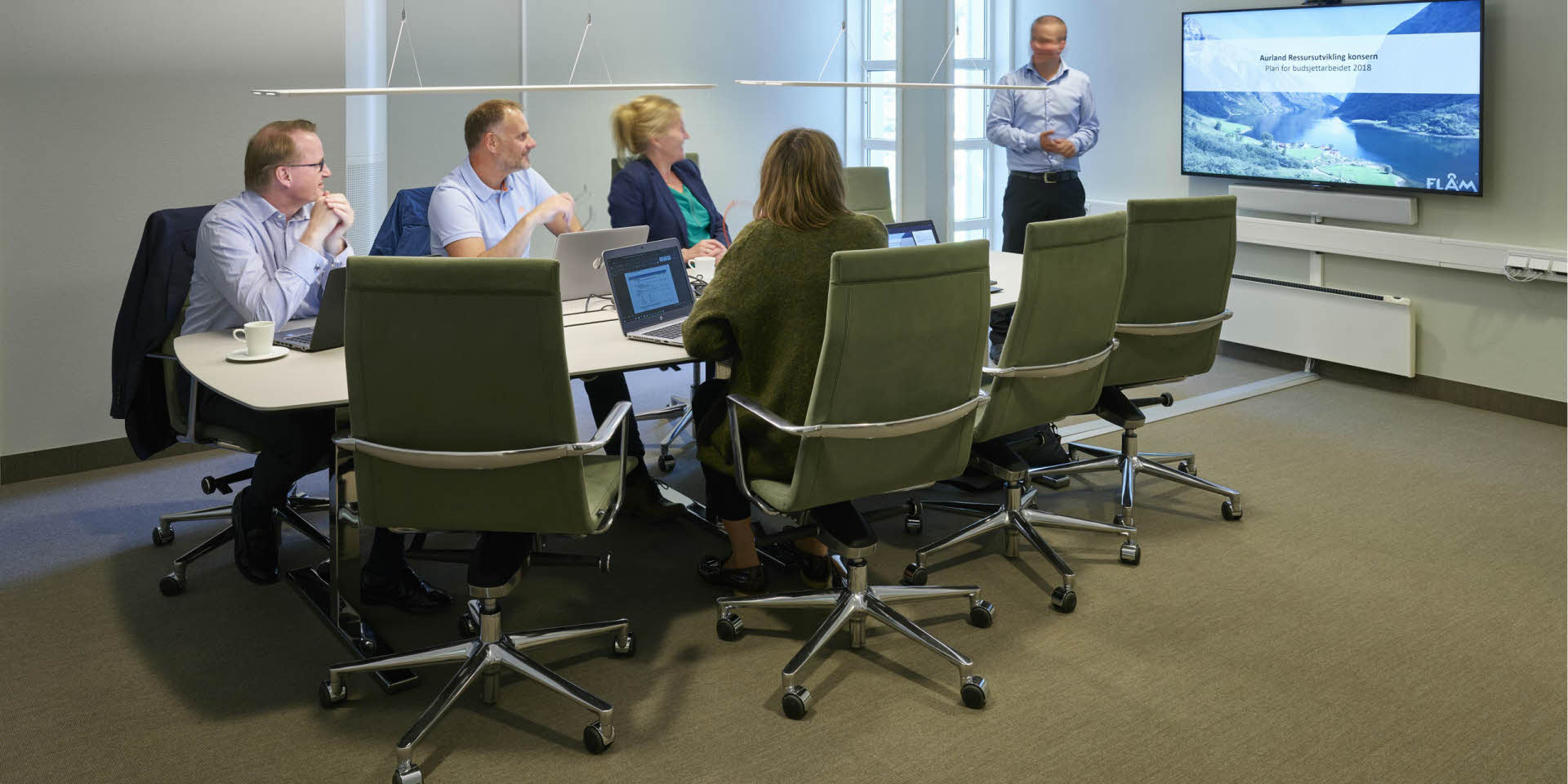Cuatro personas sentadas en torno a una mesa observan a un hombre que realiza una presentación en el hotel Fretheim