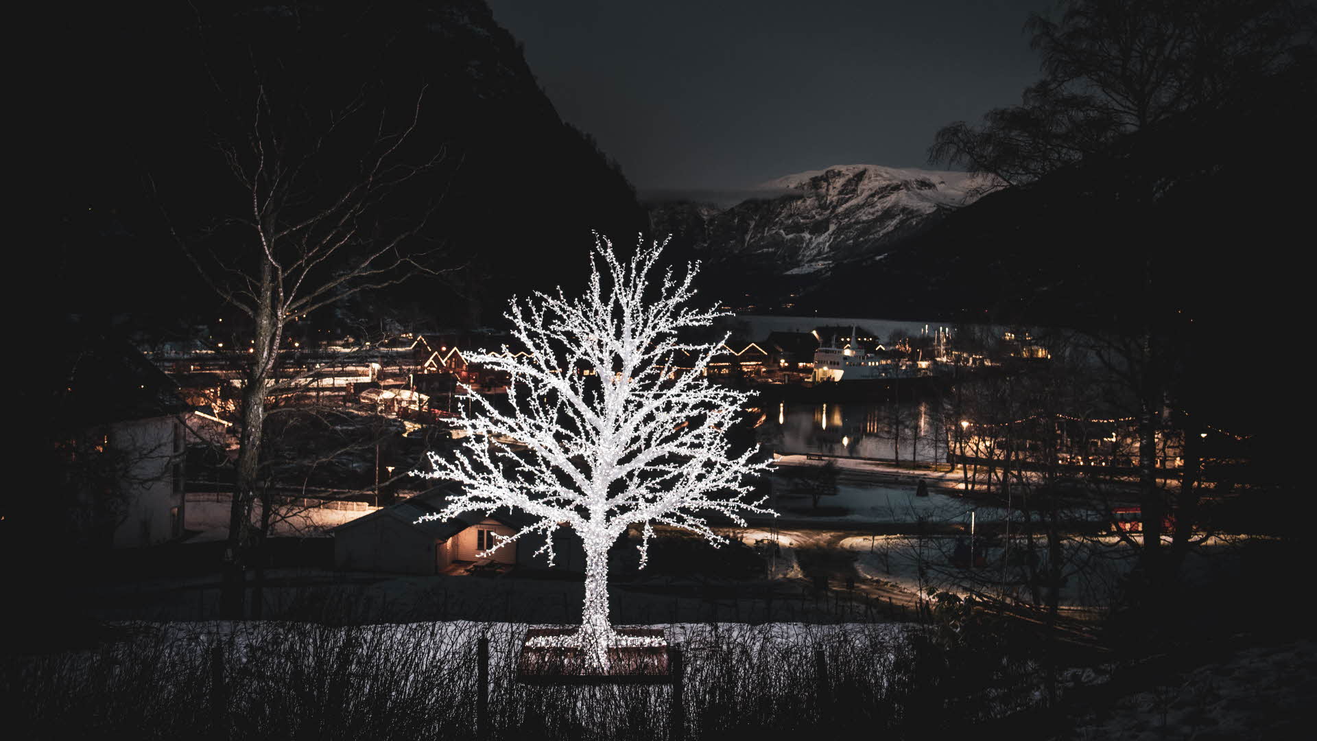 Ein silberner Lichterbaum im Vordergrund, Flåm mit Weihnachtsbeleuchtung, im Hintergrund der Fjord und die schneebedeckten Bergen. 