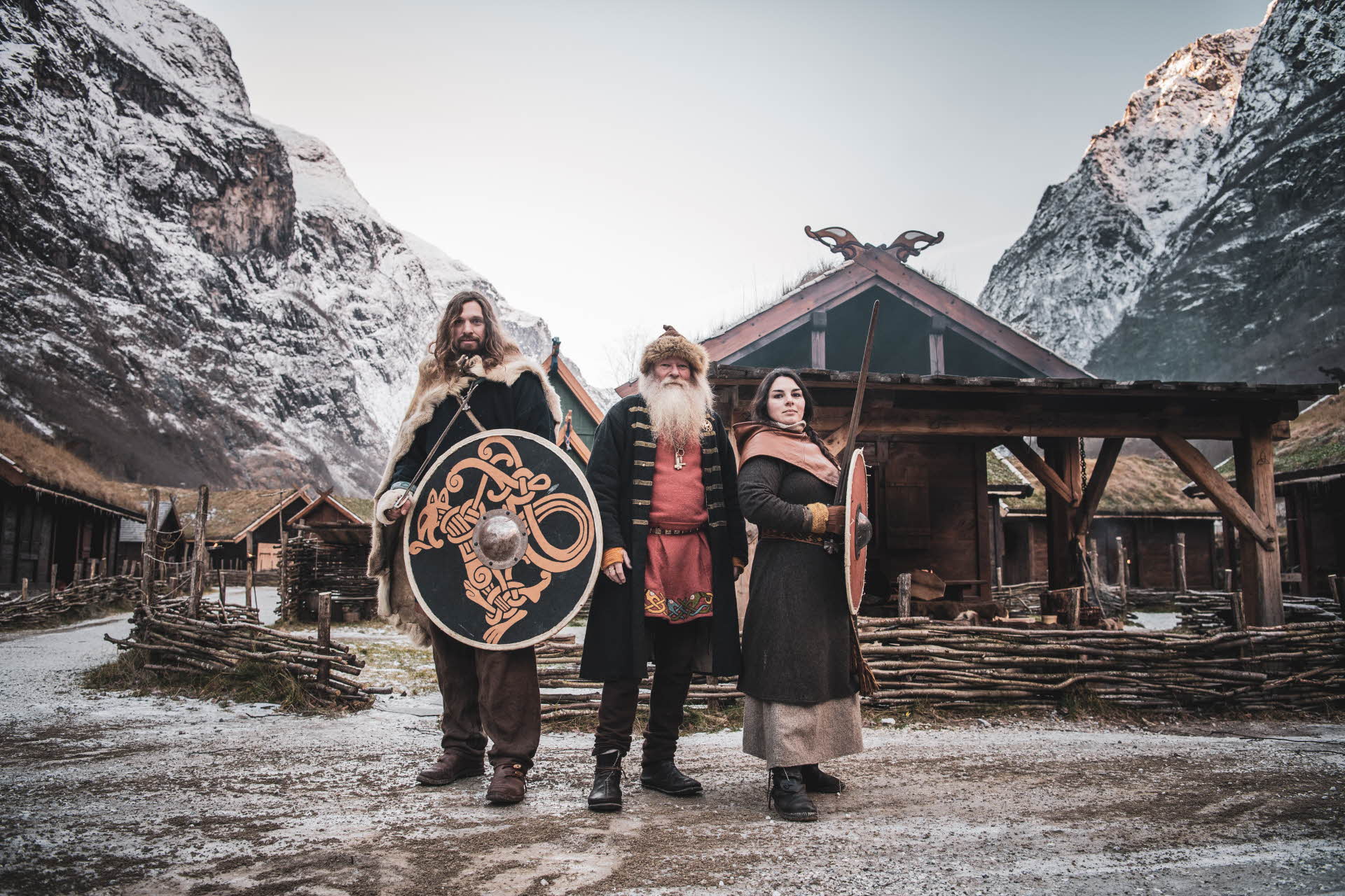 Tre vikinger foran et vikinghus og fjelltopper med nysnø i Gudvangen