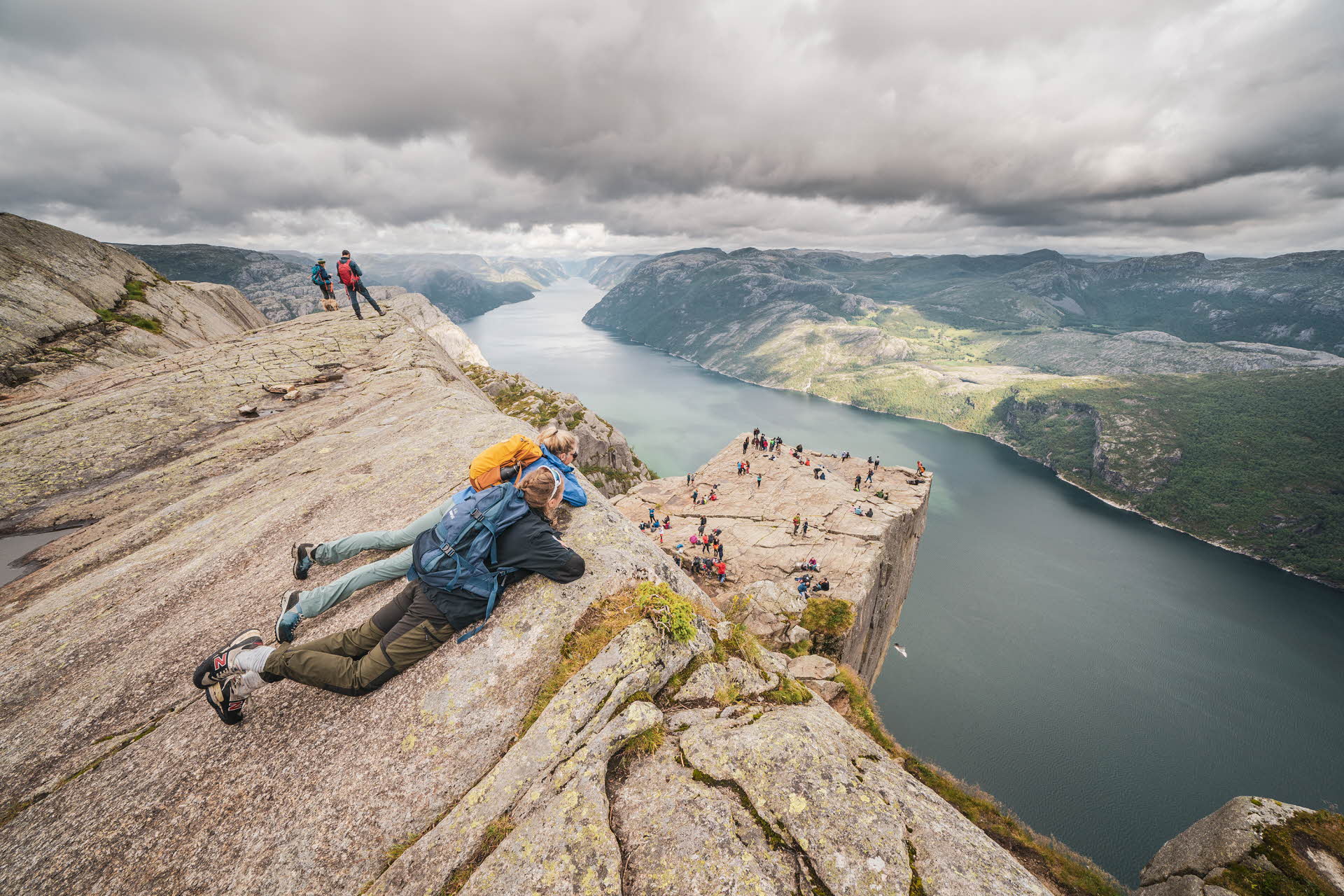 Two women lying on a mountain cliff looking down at a crowd on the Pulpit Rock and the fjord below