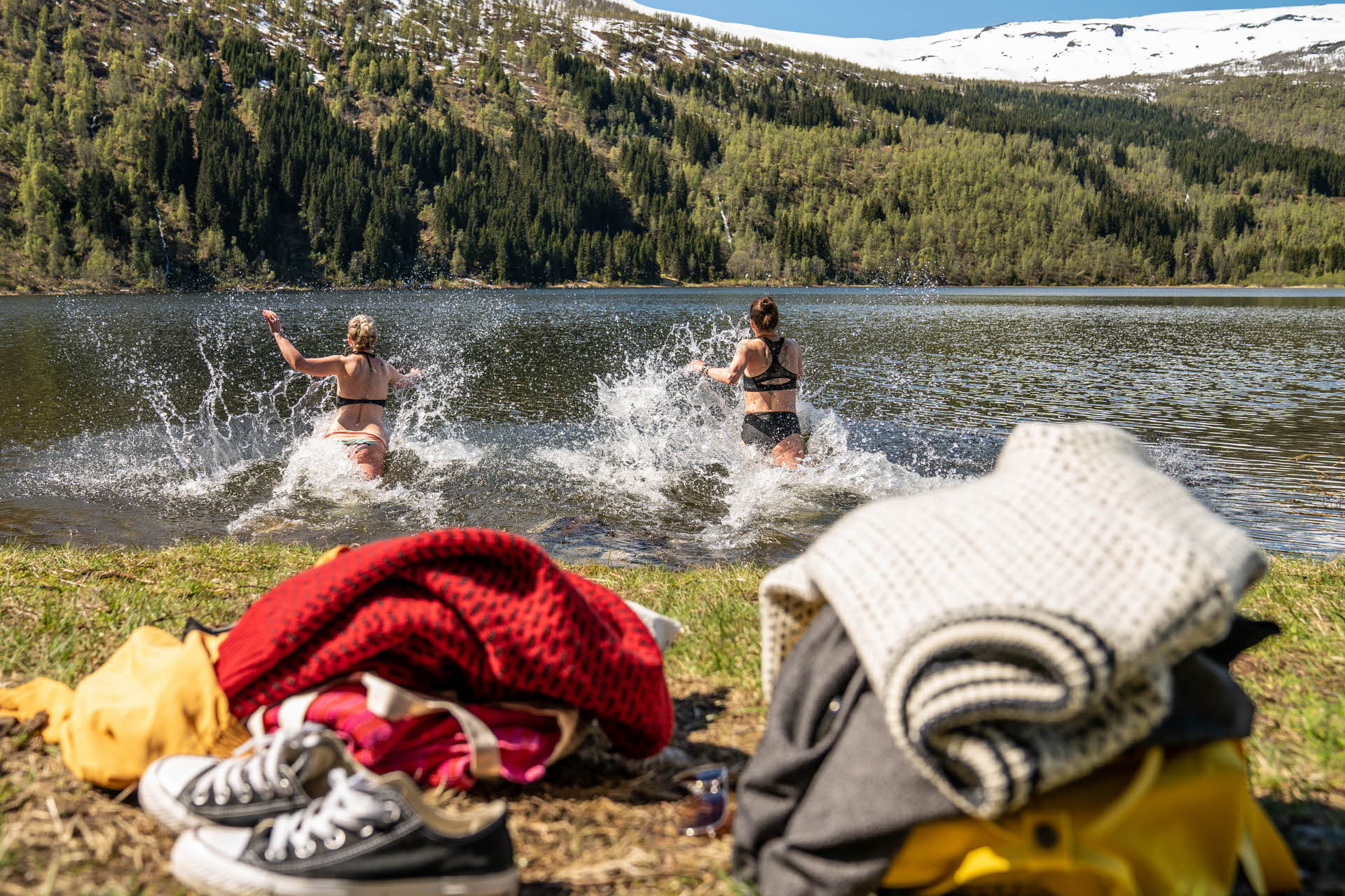 With their clothing and shoes on the ground in front, two women run into a lake with snow-capped mountains in the background. 