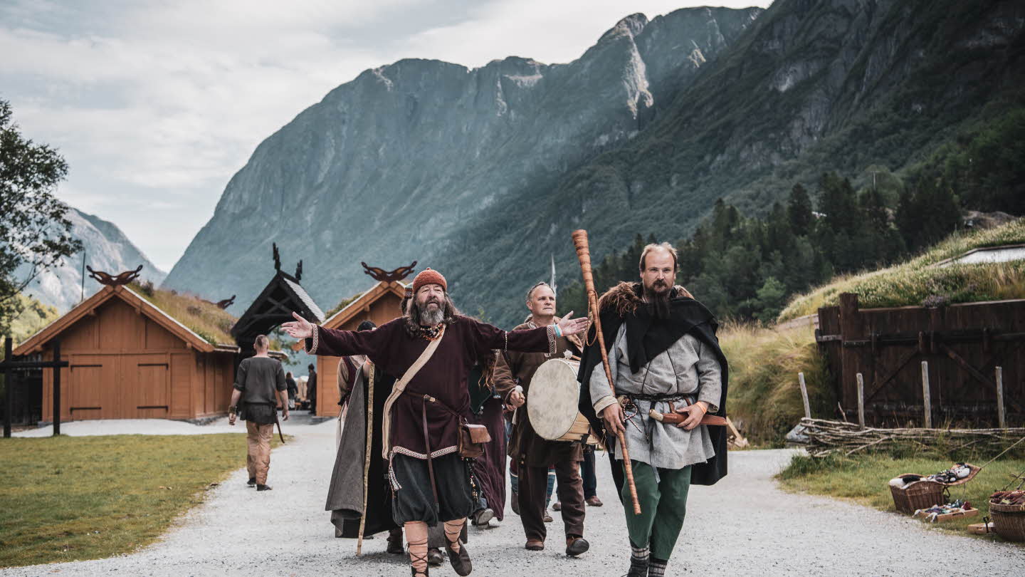 A group of men dressed as Vikings walk on a dirt road in front of Viking-like wooden houses with turf roofs and steep mountain sides