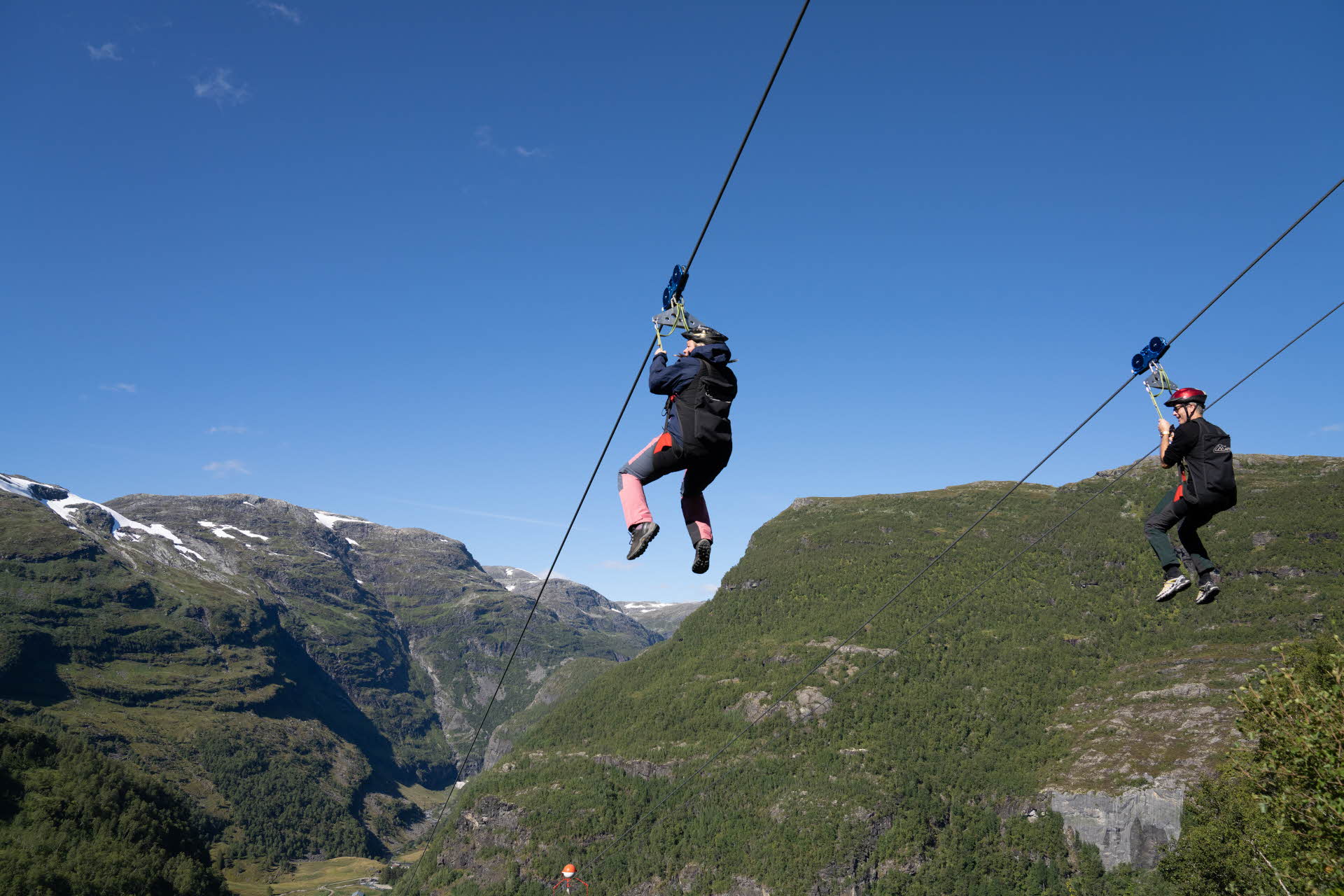 Zwei Personen hängen in einem Geschirr von der Flåm Zipline, umgeben von blauem Himmel und grünen Bergen. 