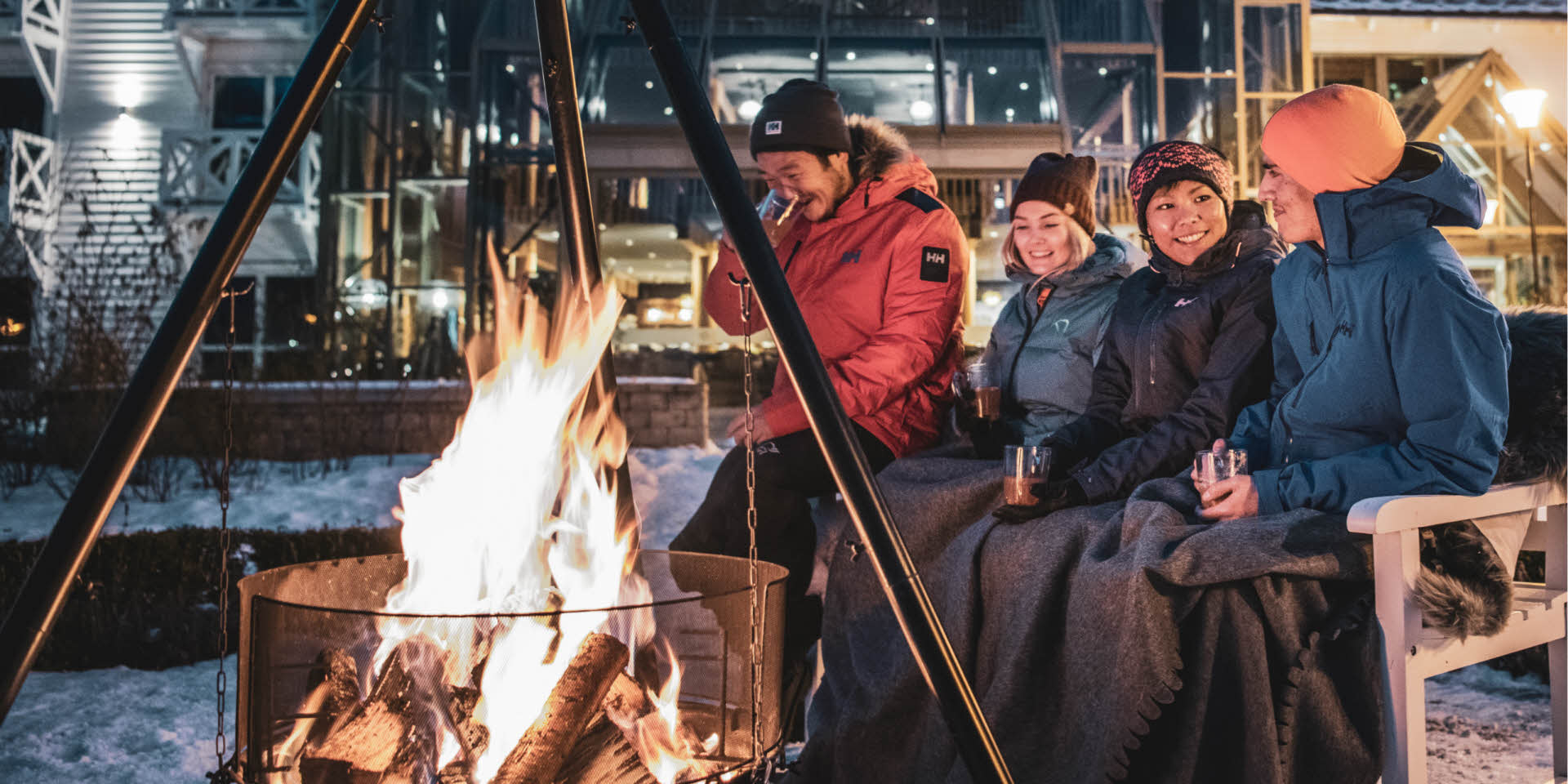 Four people sitting on a bench by the lit bonfire outside Fretheim Hotel, while drinking gløgg and smiling