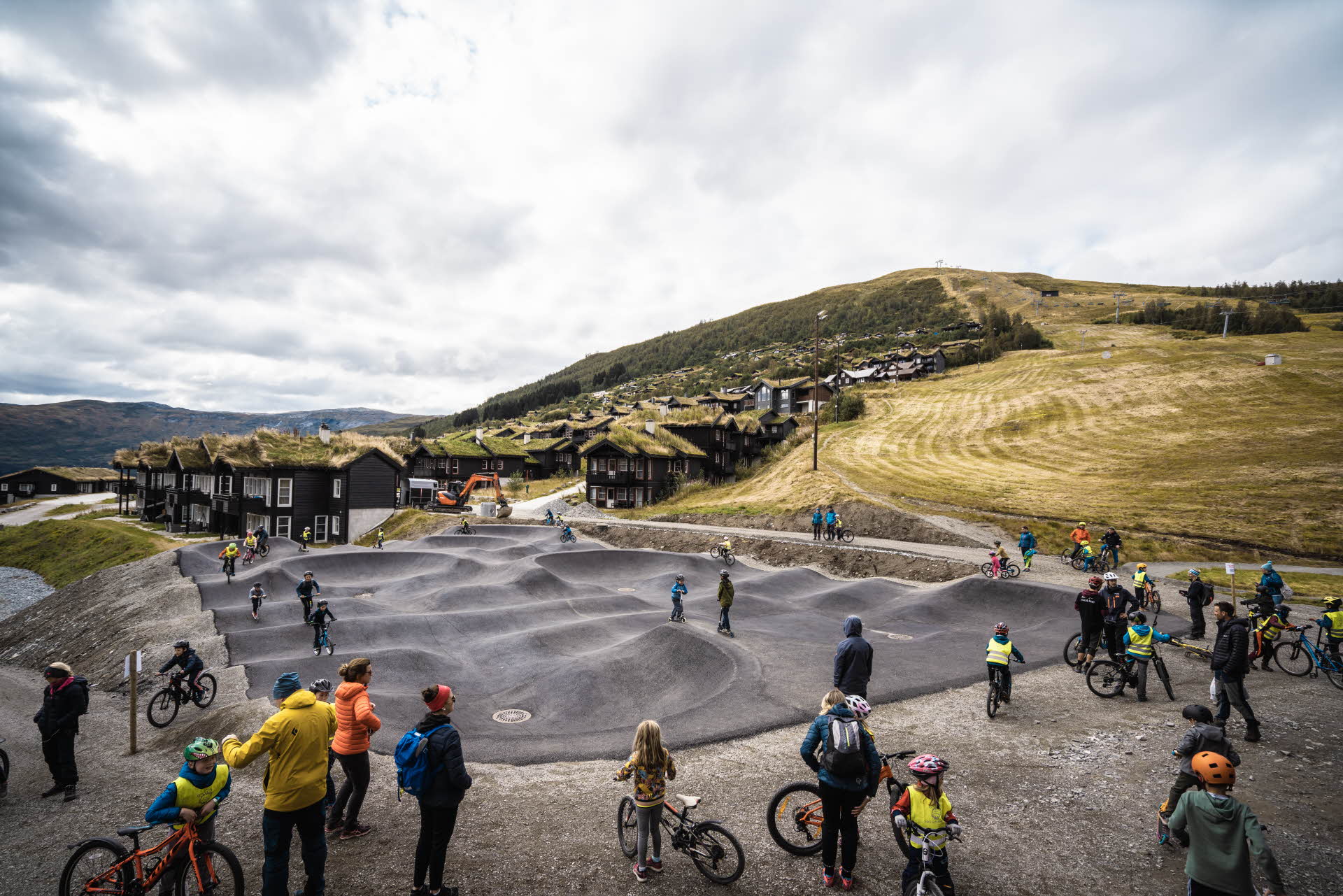 Varios niños y adultos en bici y de pie alrededor del circuito de pumptrack, en Myrkdalen Fjellandsby